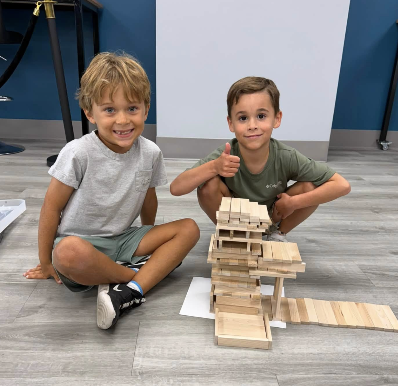 Two boys sitting on the floor next to a small wooden architectural model, smiling at the camera. One boy is giving a thumbs-up.
