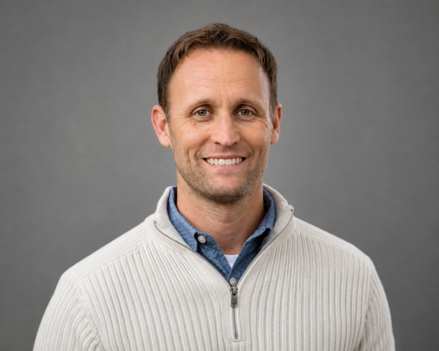 A smiling man with short brown hair, wearing a light-colored zip-up sweater over a blue collared shirt, standing against a plain gray background.