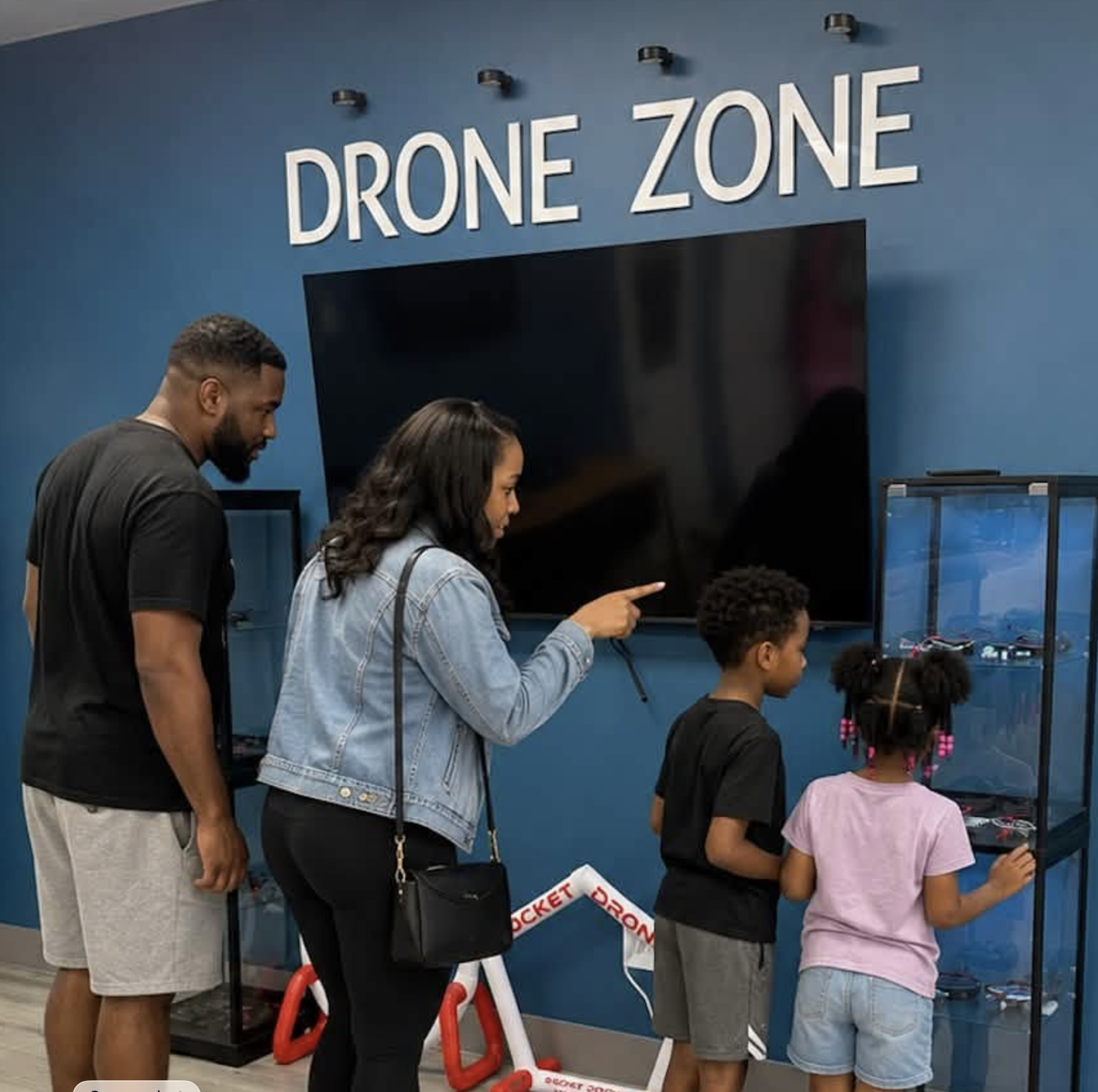 People standing in line at a drone zone, with a large monitor and a drone display case. Signage reads 'Drone Zone' and a barrier with 'Rocket Drop' is visible.