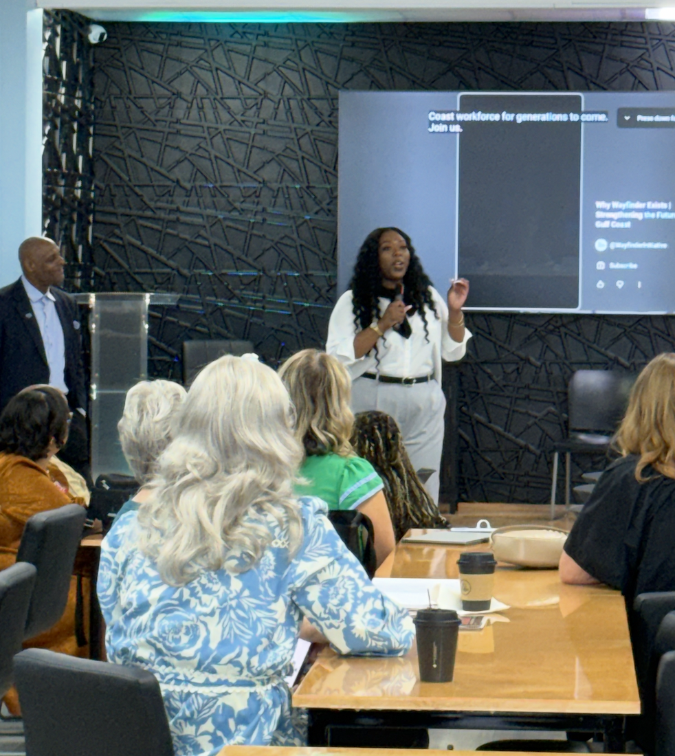 A woman stands in front of a large screen giving a presentation to a seated audience in a conference room. A man stands to her side. The audience includes women of different ages and hair colors, some with coffee cups, paying attention to the speaker.
