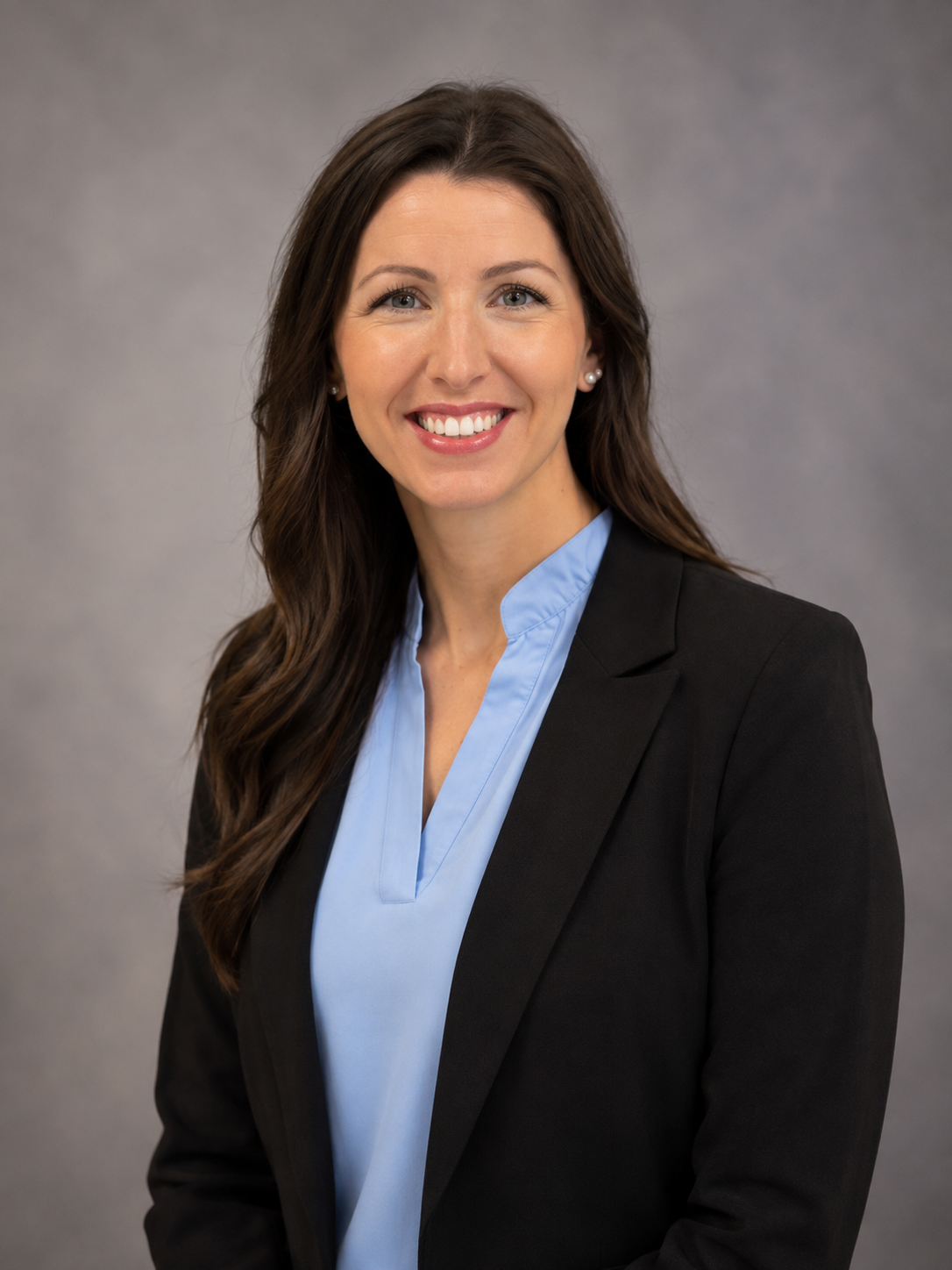Professional woman with long brown hair, wearing a black blazer and light blue blouse, smiling in a studio portrait.