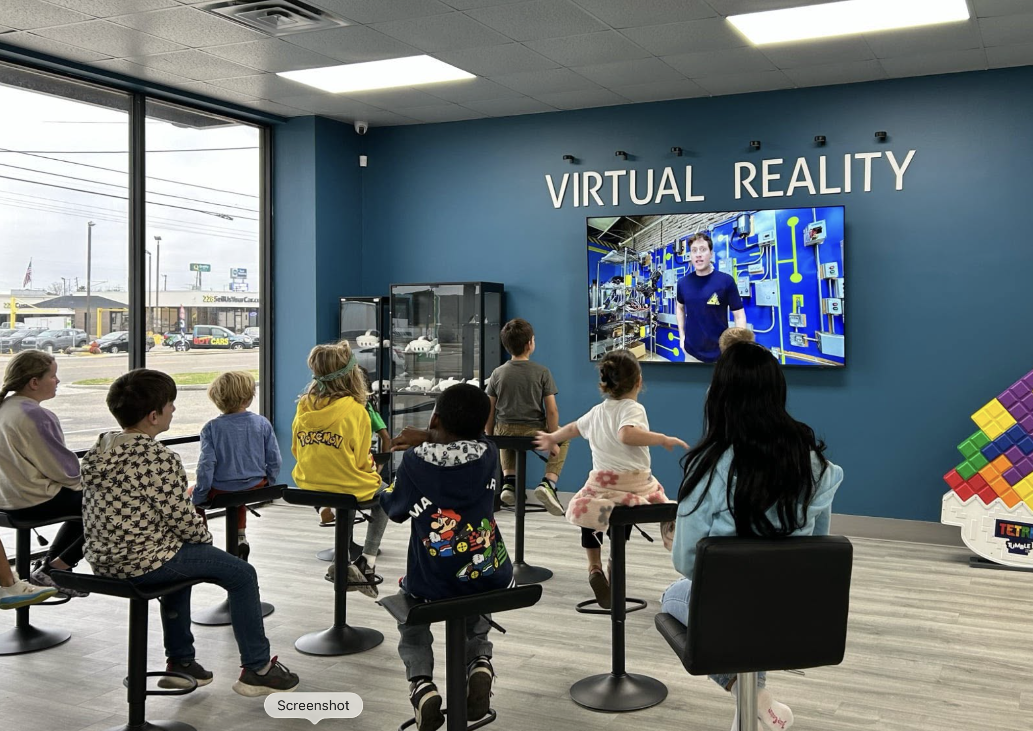Children attending a presentation about virtual reality in a store or showroom with a large screen displaying a man explaining VR technology.