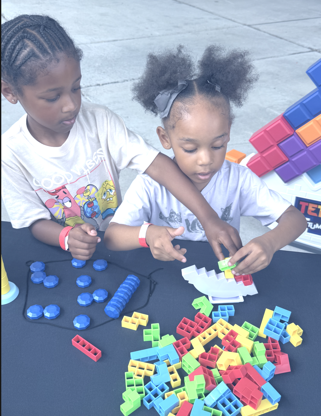 Two young girls playing with colorful building blocks and tokens at a table, outdoors.