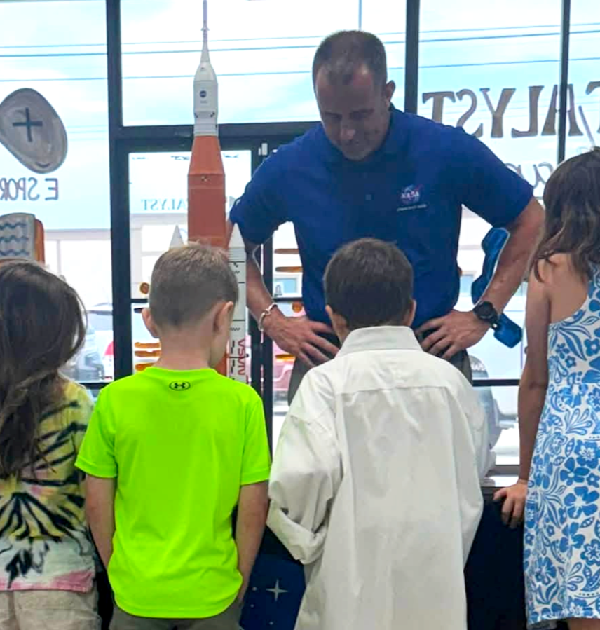 A man in a NASA shirt demonstrating a rocket model to children inside a store with large windows.