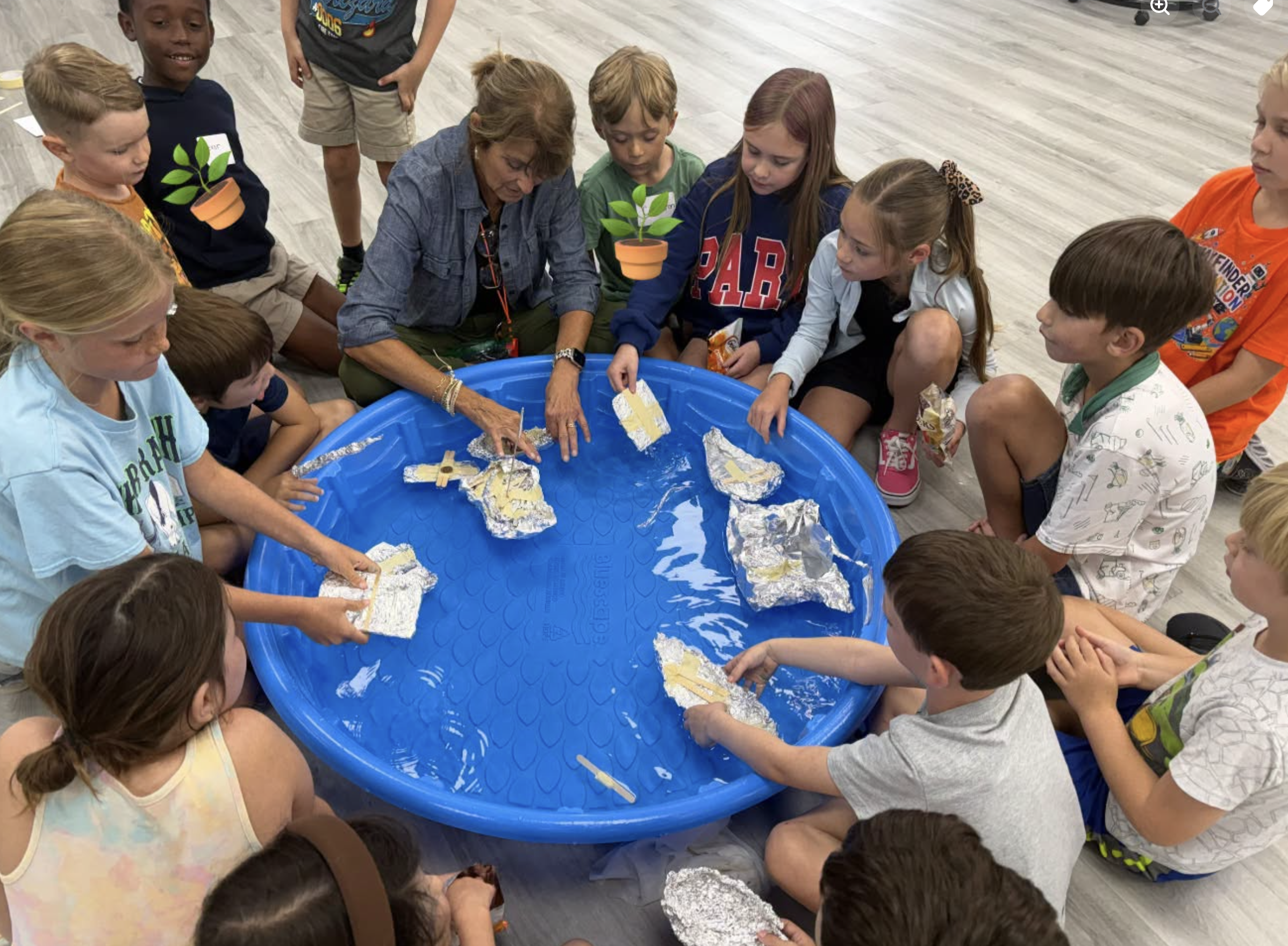 A group of children gathered around an adult woman at a round, blue water table, engaging in a hands-on activity with aluminum foil and paper towel rolls in an indoor classroom or activity space.