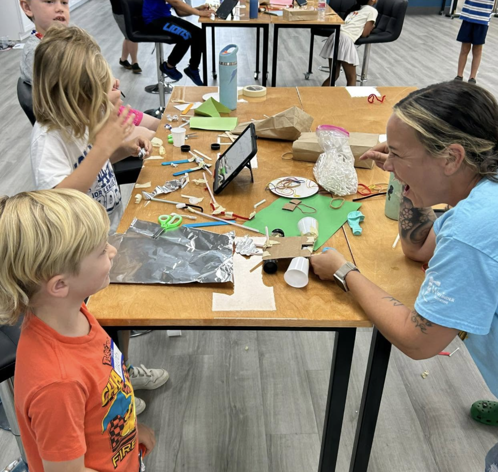 Children and an adult at a table engaged in a science or craft activity with various materials, papers, scissors, and supplies scattered on the table in a classroom or activity setting.