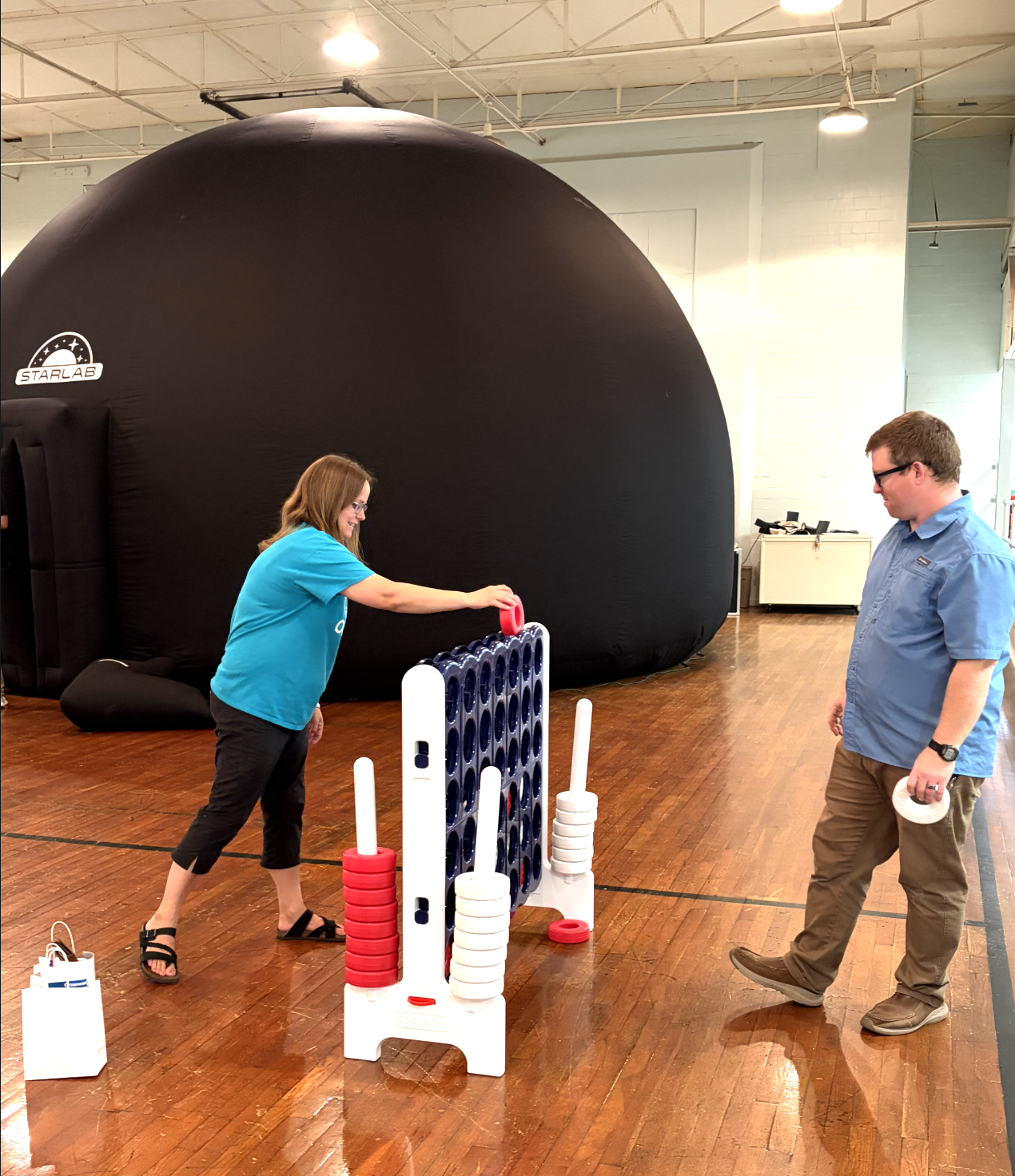 Two people playing a giant outdoor connect four game inside a room with wooden floors and high ceilings, with a large black inflatable object in the background.