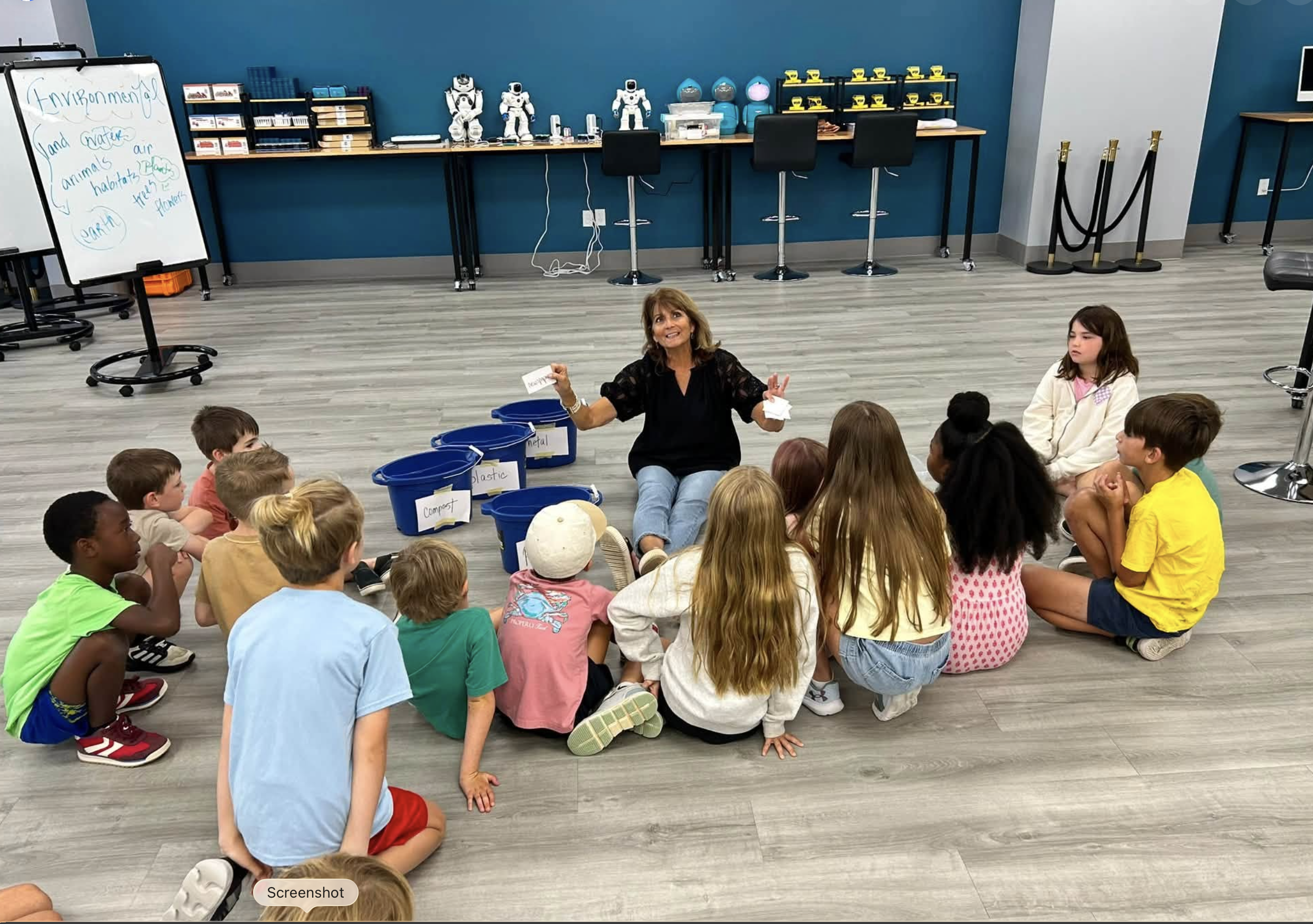 A woman sitting cross-legged on the floor, surrounded by children, leading a discussion in a classroom or workshop setting. Behind her are educational bins labeled with words like 'plastic,' 'metal,' and 'compost,' and a whiteboard with notes about the environment and habitats. The background features a table with robotics or science equipment and a row of robots.