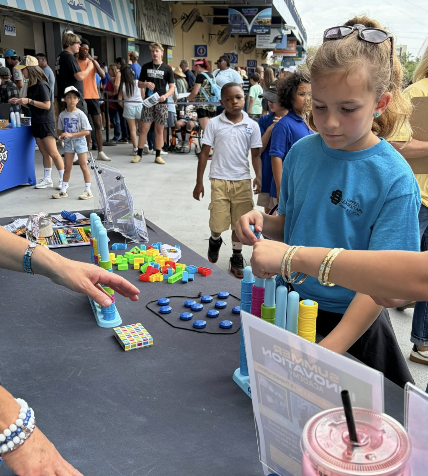 Children playing a board game at an outdoor event with a crowd in the background.