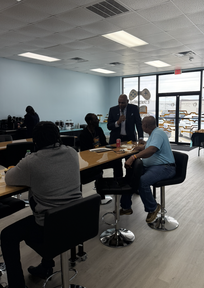 Four men sitting and standing around a high table in a room with a coffee station in the background, engaging in conversation.