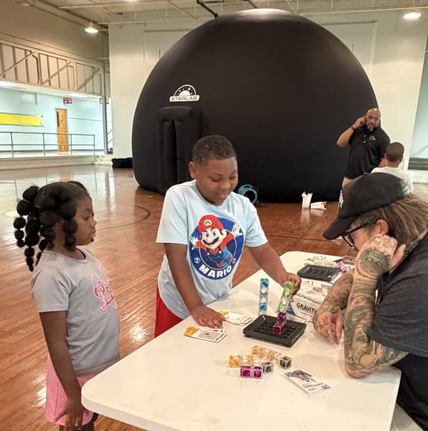 Children and a man playing a board game called Gravitation at a table inside a building with a wooden floor, with a large black inflatable dome in the background.