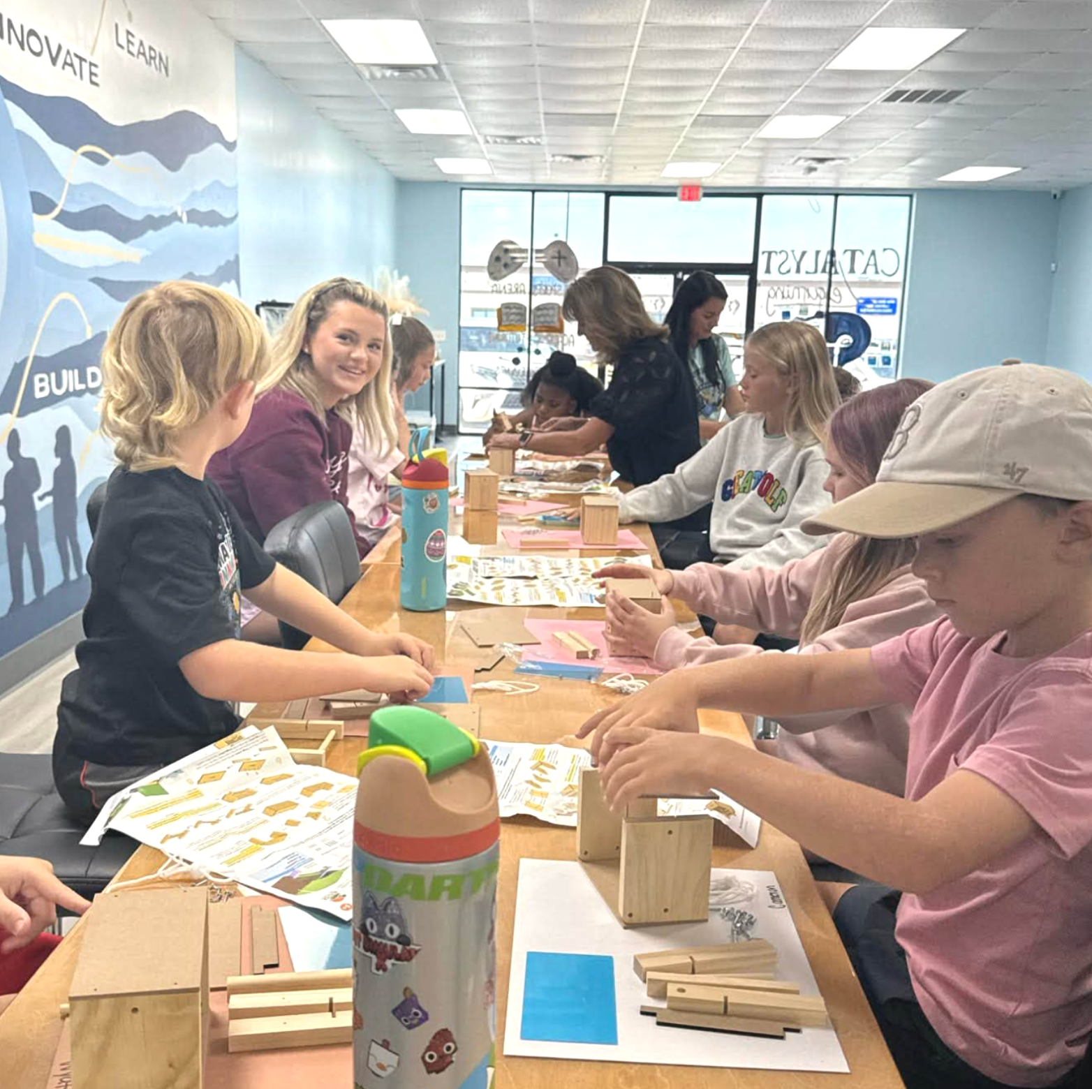 Children and adults seated at a table engaging in a woodworking activity inside a room with light blue walls and large windows.