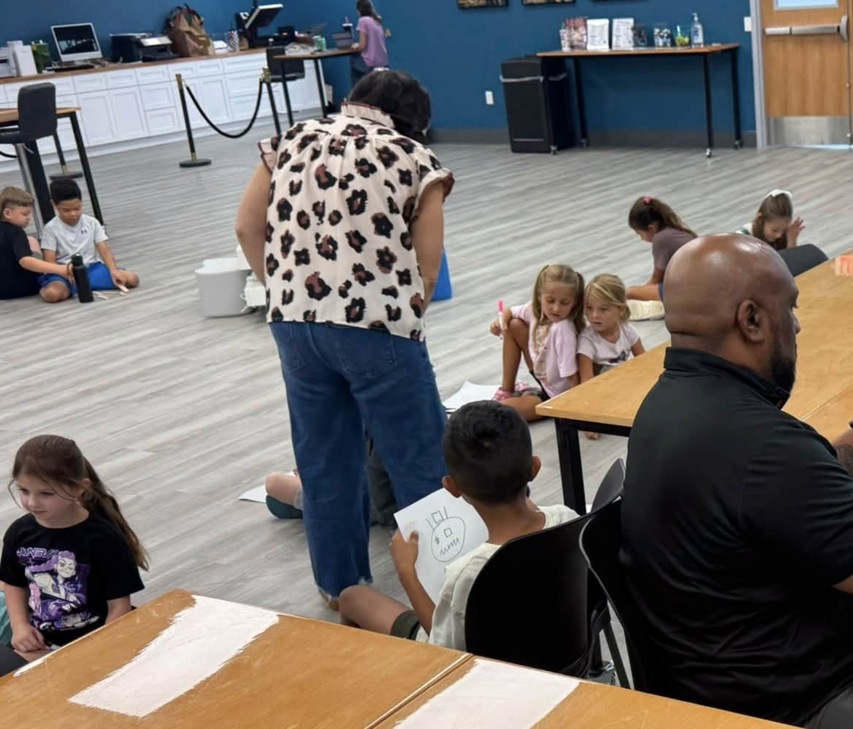 Children sitting on the floor and at tables in a room with blue walls, engaging in drawing activities, supervised by adults. Some children are focusing on their artwork, while others look around or chat.