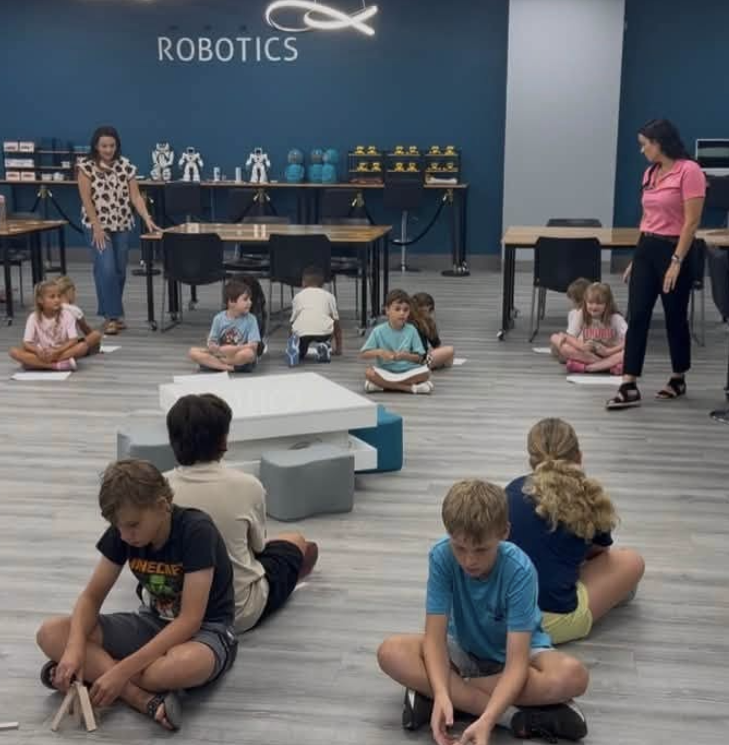 Kids sitting on the floor in a robotics classroom with a blue wall that says "ROBOTSICS" and robot models on display in the background.