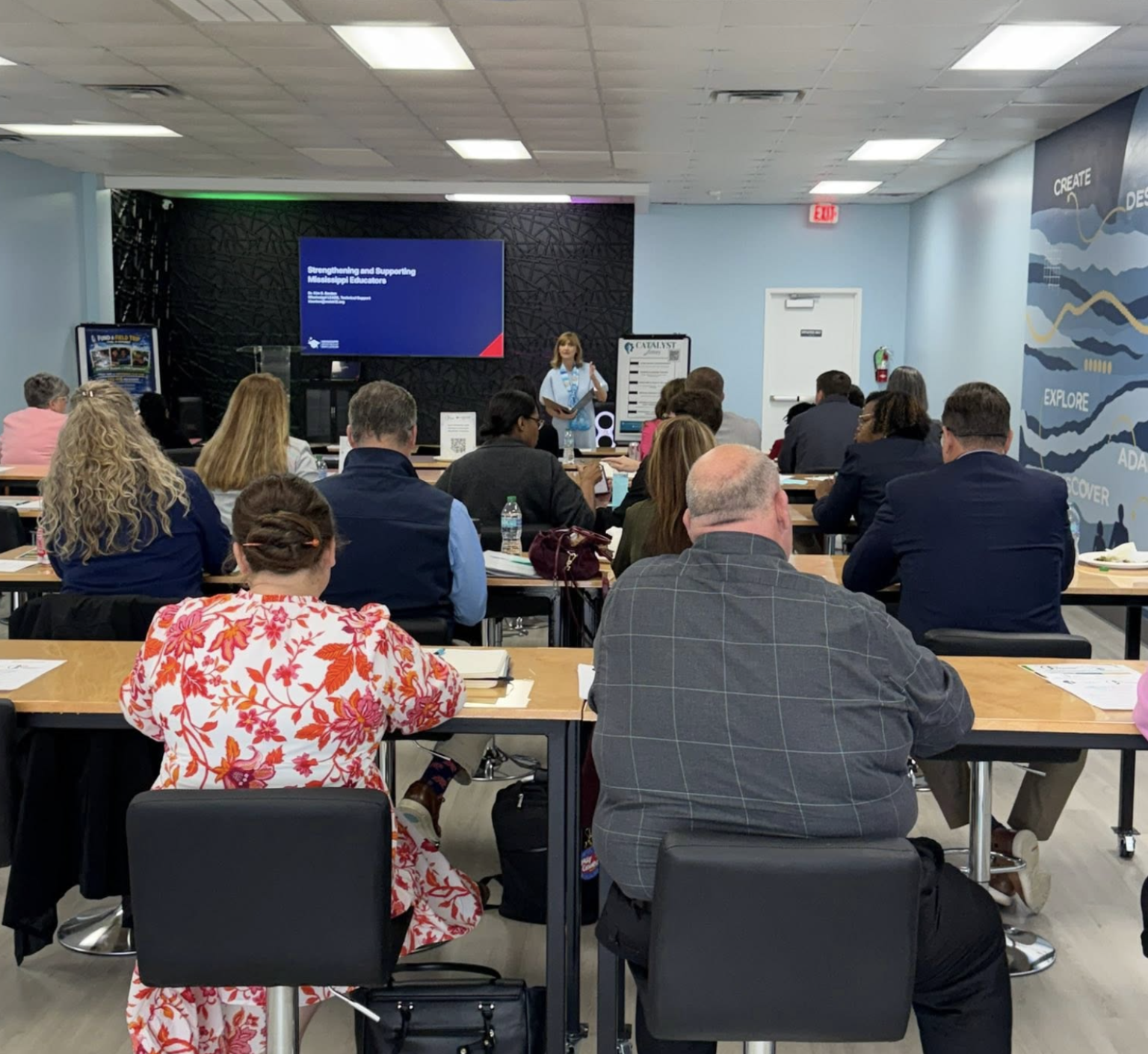 A group of people attending a conference or seminar in a room with a speaker presenting at the front. The attendees are seated at tables, facing the speaker and a large screen displaying a presentation slide.