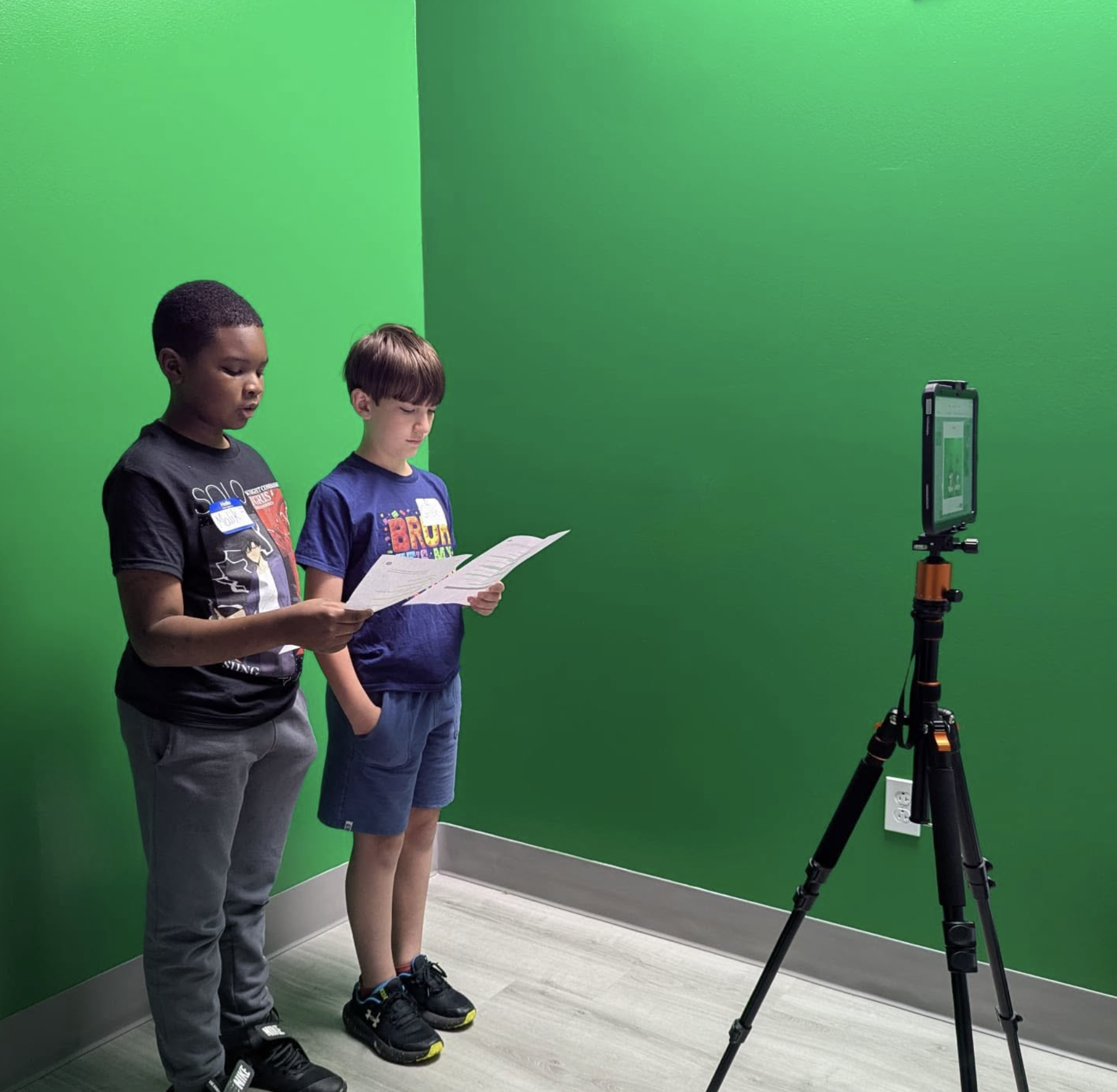Two boys standing side by side in front of a green screen, reading papers, with a camera on a tripod recording their actions.