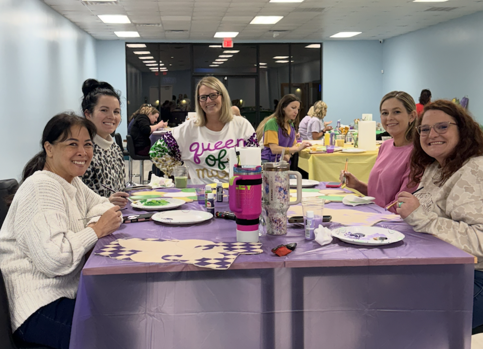 Group of seven women sitting and standing around a table painting at a craft party in a room with pastel blue walls and a large window behind them, with art supplies and drinks on the table.