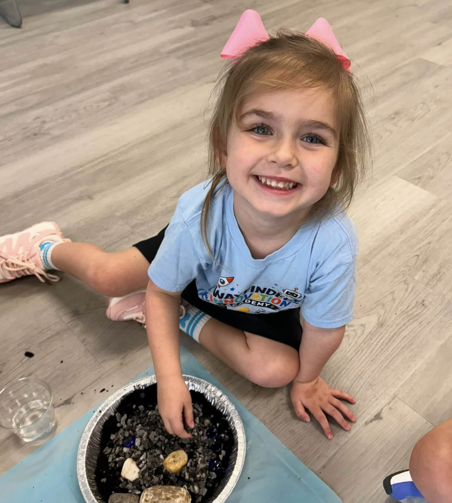 A young girl with light brown hair, blue eyes, and a big smile, sitting on the floor in a casual setting. She is wearing a blue T-shirt with colorful text, pink sneakers, and has pink paper cones in her hair resembling bunny ears. She is reaching into a large container filled with dark stones and some shells, with a few glasses and a blue cloth on the floor nearby.