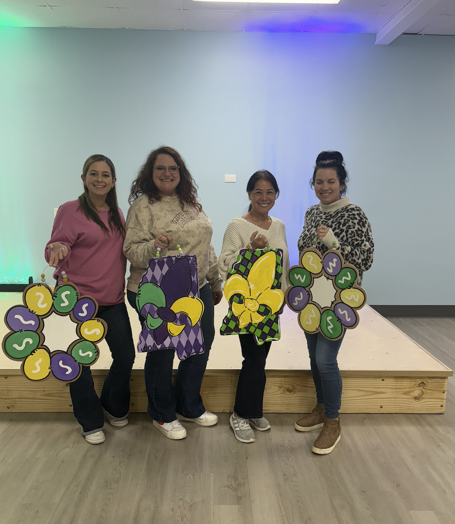 Four women standing in front of a light blue wall, each holding decorated boards with candy-themed designs, smiling at the camera.