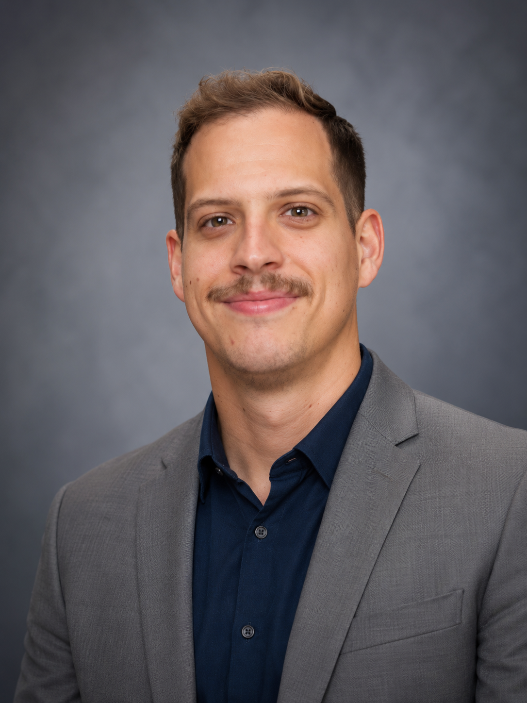 Headshot of a young man with short brown hair, a light mustache, and wearing a light gray suit jacket over a dark blue shirt, smiling against a gray background.
