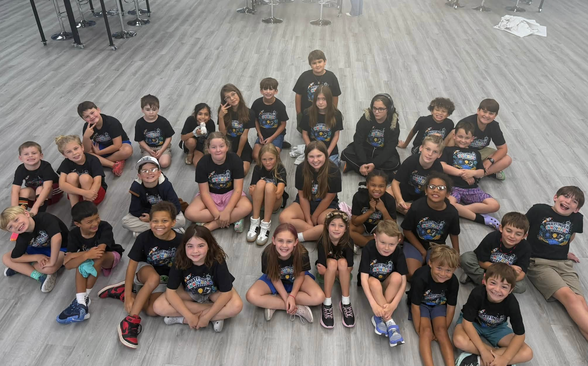 A group of children sitting on the floor in a large room, all wearing black t-shirts with a colorful logo. The children appear happy and are posing for the photo, some smiling and others making playful gestures.