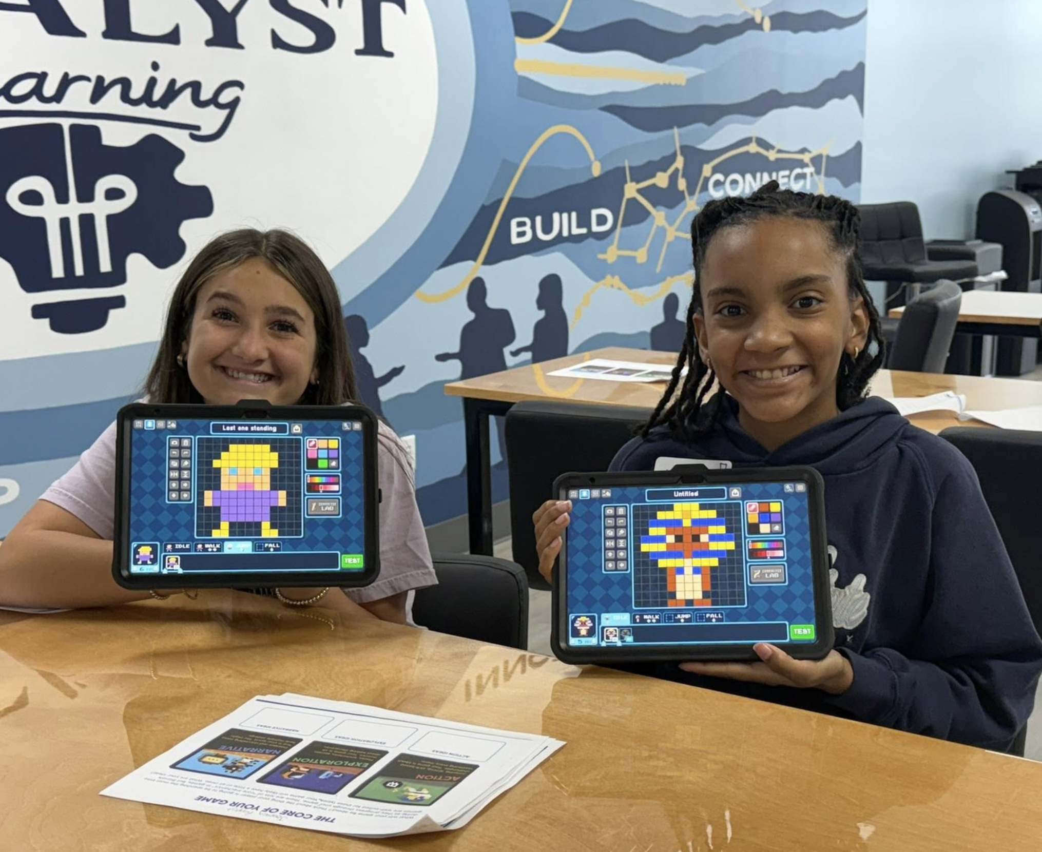 Two young girls are sitting at a wooden table, smiling and holding tablets displaying pixel art images of people. Behind them, a blue wall features motivational words 'BUILD' and 'CONNECT' alongside a graphic of silhouettes and a line graph. There are printed sheets on the table and office chairs in the background.