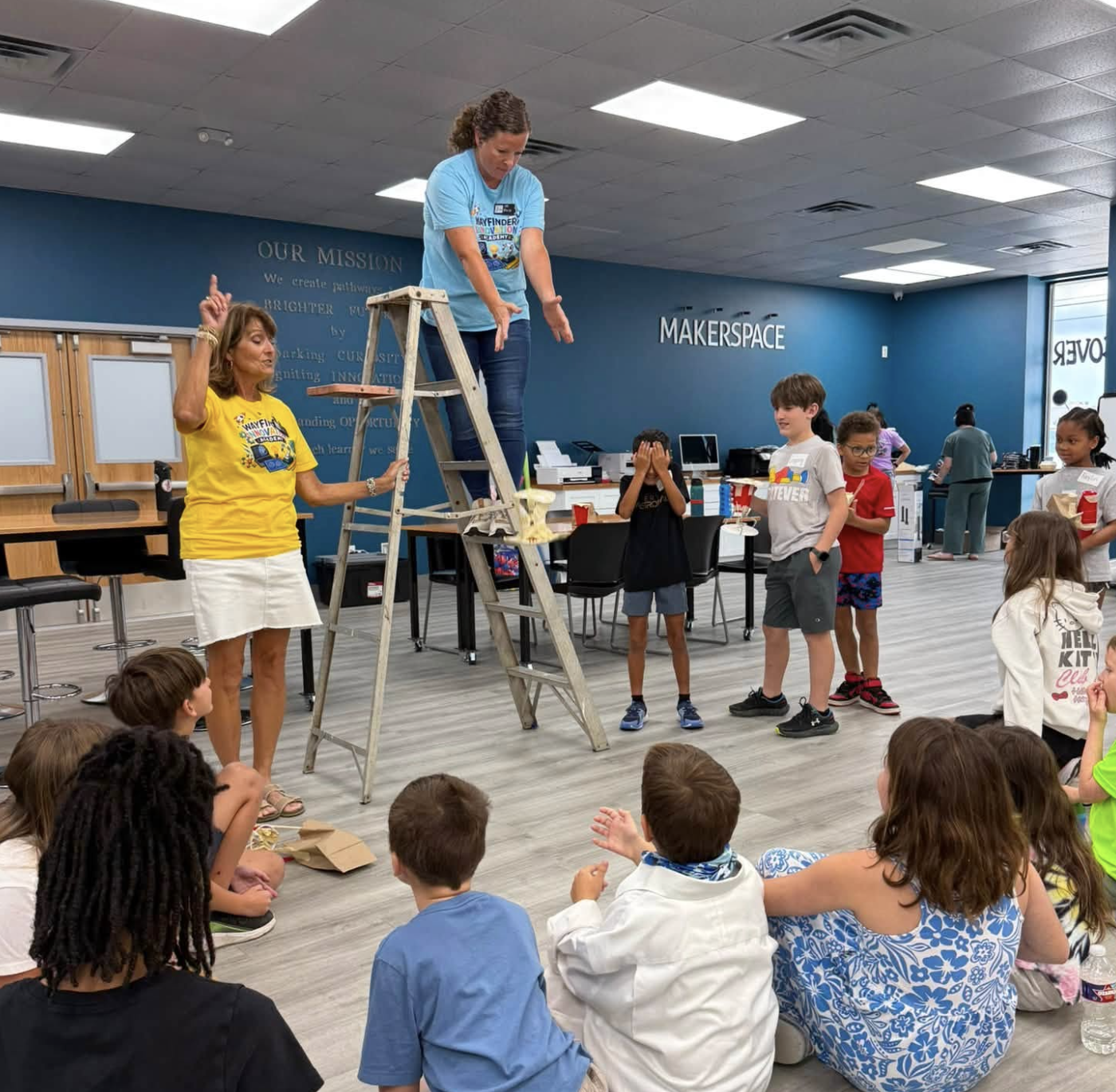 A group of children participating in a science activity while seated and standing in a makerspace classroom. Two women, one dressed in yellow, facilitate the activity involving a ladder and a substance on top. The background features a blue wall with the words "Makerspace" and "Our Mission." Children are watching and some are covering their eyes or raising their hands.