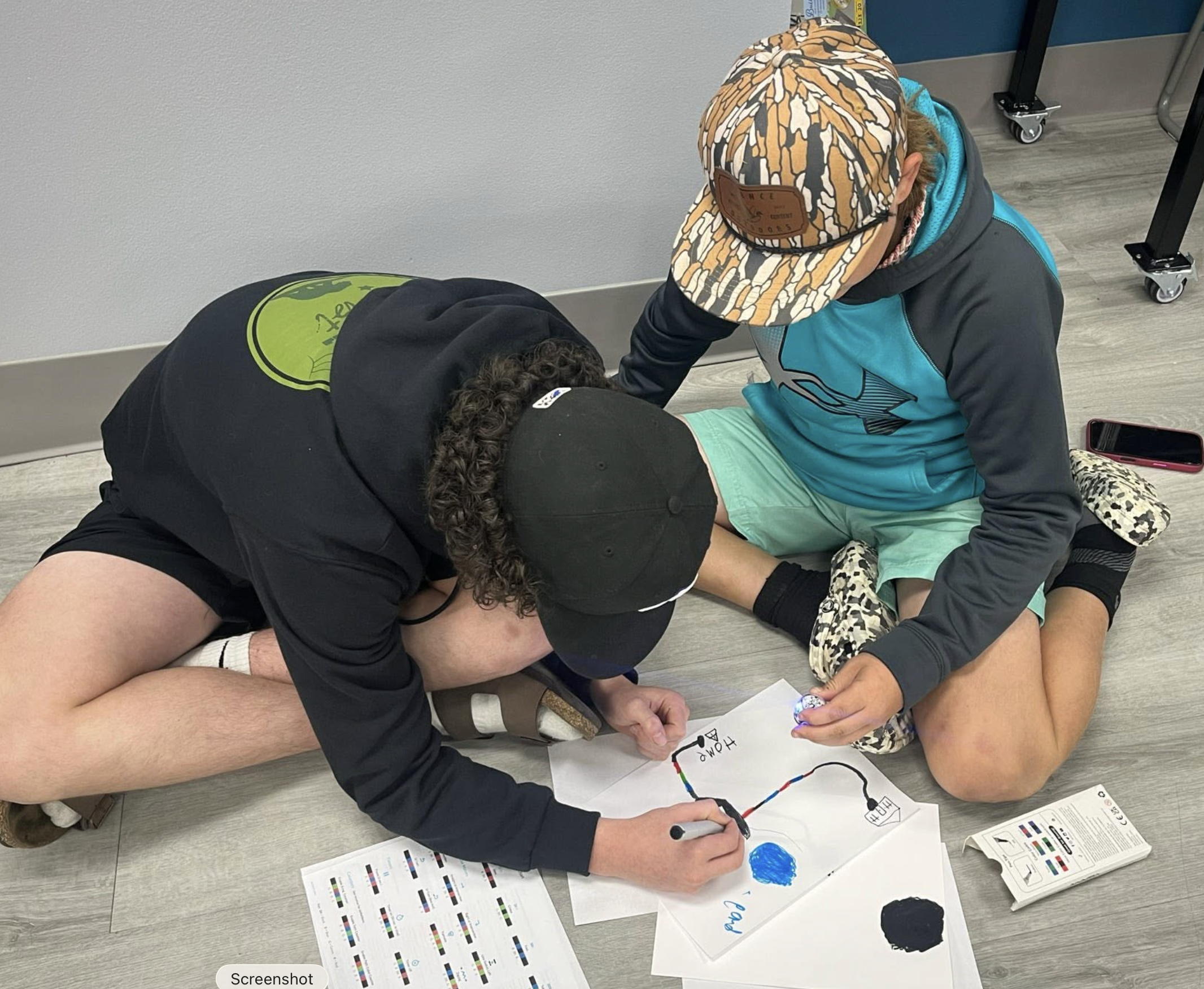 Two children are sitting on the floor, one is lying down and another is kneeling, working on a project involving a colorful diagram and written notes. The diagram includes a curved line and various labeled components, with papers and markers scattered around.