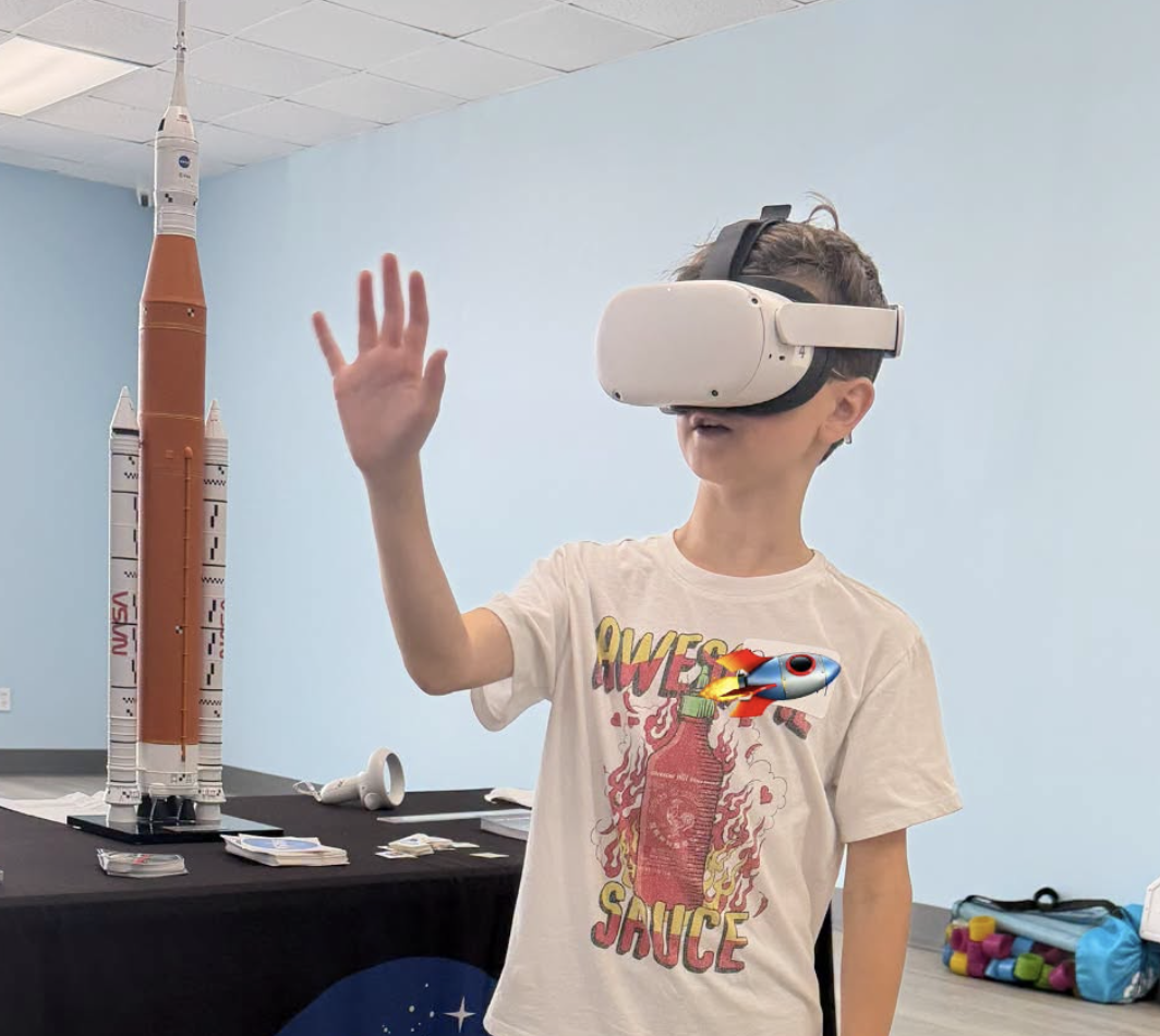 Child wearing a virtual reality headset and reaching out with hand, standing next to a model rocket on display in a room with a black table and blue walls.