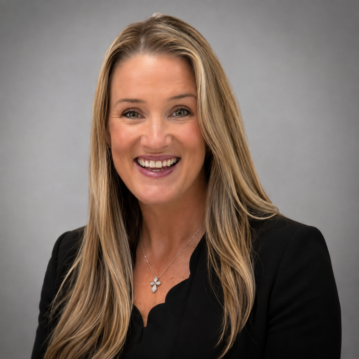 A smiling woman with long blonde hair, wearing a black blazer and a flower pendant necklace, against a plain grey background.