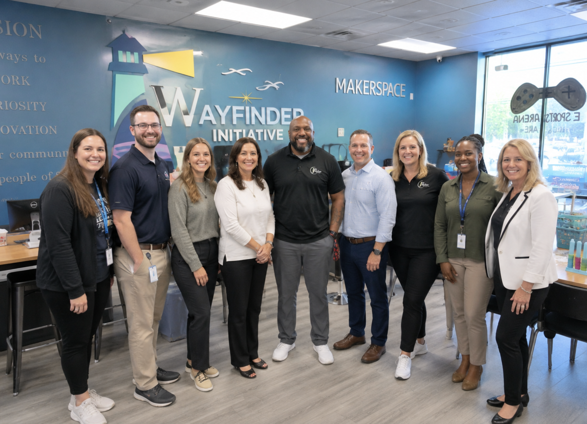 Group of nine diverse smiling people standing together inside a community space with a blue wall that reads 'WAYFINDER INITIATIVE' and includes a lighthouse design, large windows, and some tables and chairs.