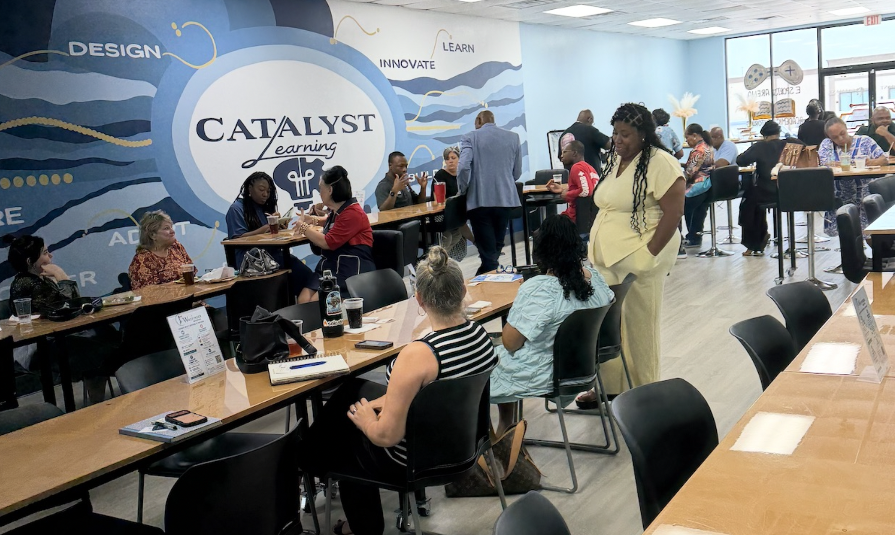 People sitting and standing in a modern, brightly lit classroom or workshop space with tables, chairs, and a vibrant mural on the wall that reads 'Catalyst Learning' with words like 'Design,' 'Learn,' 'Innovate,' and 'Adapt.'