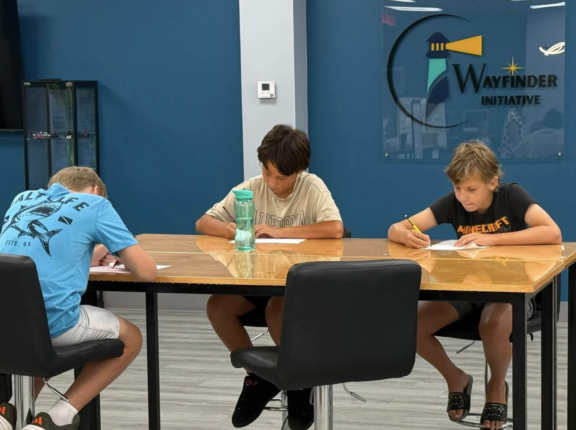 Three boys sitting at a modern wooden table in a room with blue walls and a logo of the Wayfinder Initiative on the wall. The boys are focused, writing or drawing on papers in front of them. One is wearing a blue shirt, another a beige shirt, and the third a black shirt with a gaming logo.