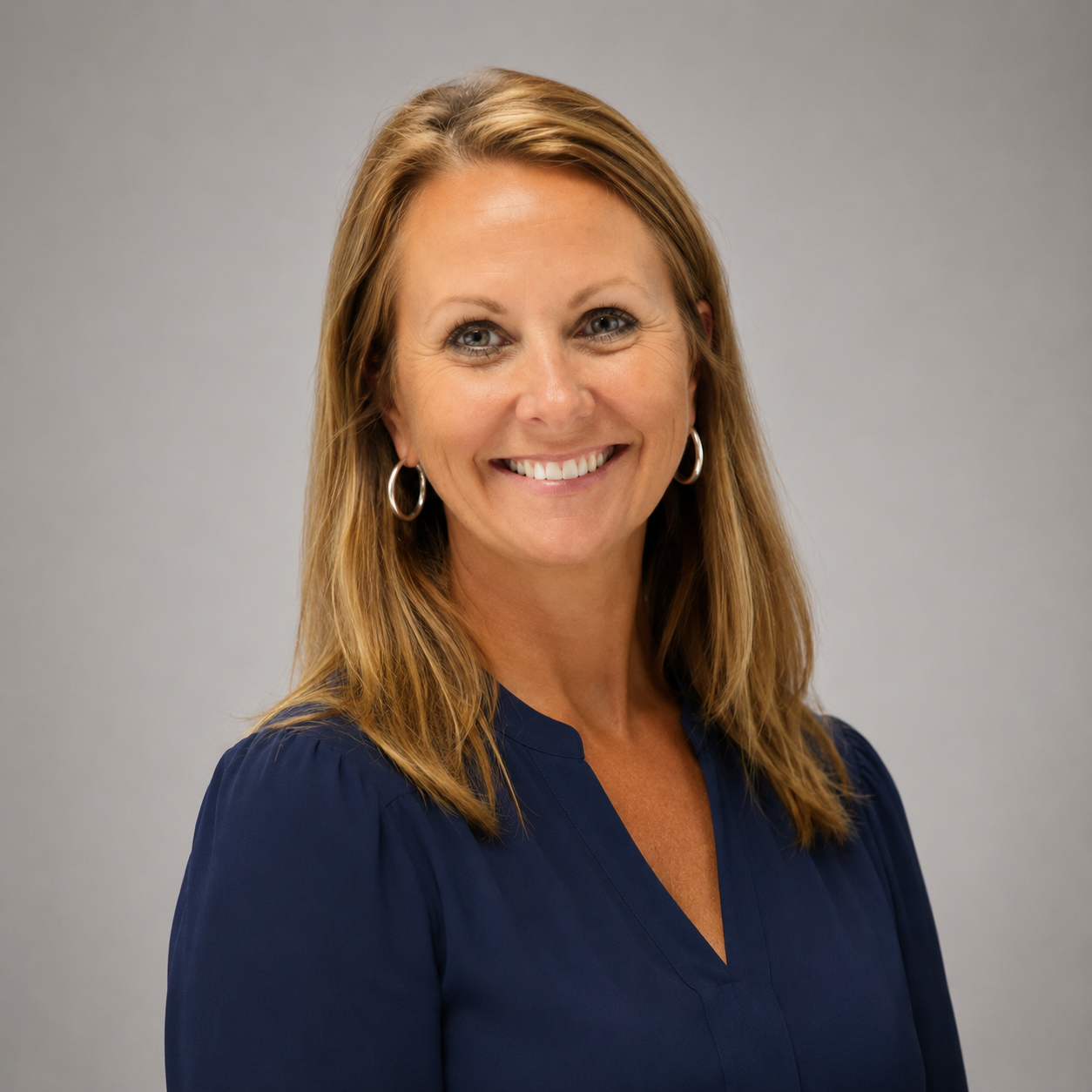 A smiling woman with shoulder-length light brown hair, wearing hoop earrings and a navy blue blouse, posing against a plain gray background.