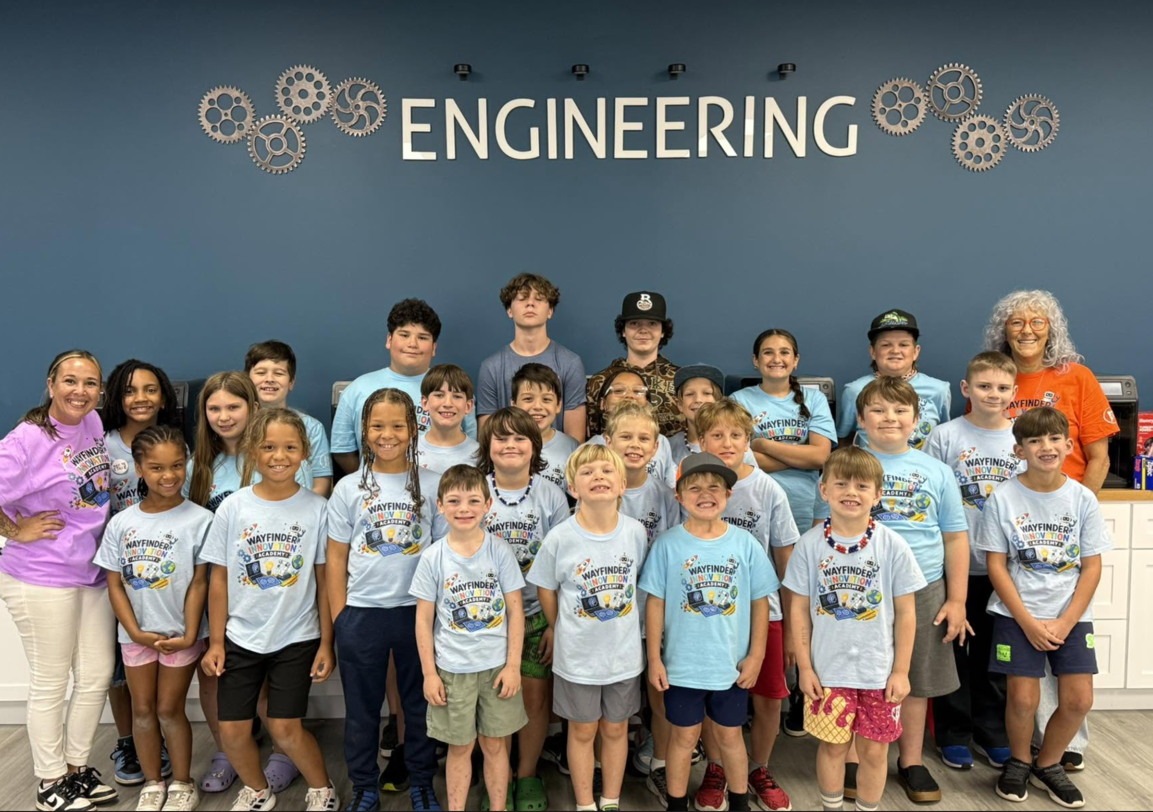 Group of children and their teacher in front of a blue wall with large white letters reading 'ENGINEERING' and decorative gears.