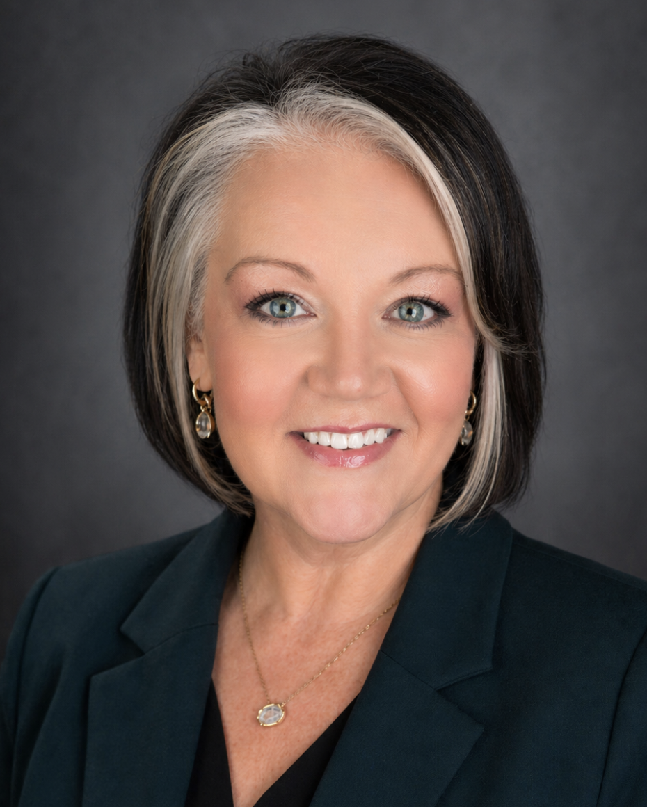 A woman with short dark hair with blonde streaks, wearing a dark blazer, gold jewelry, smiling against a dark background.