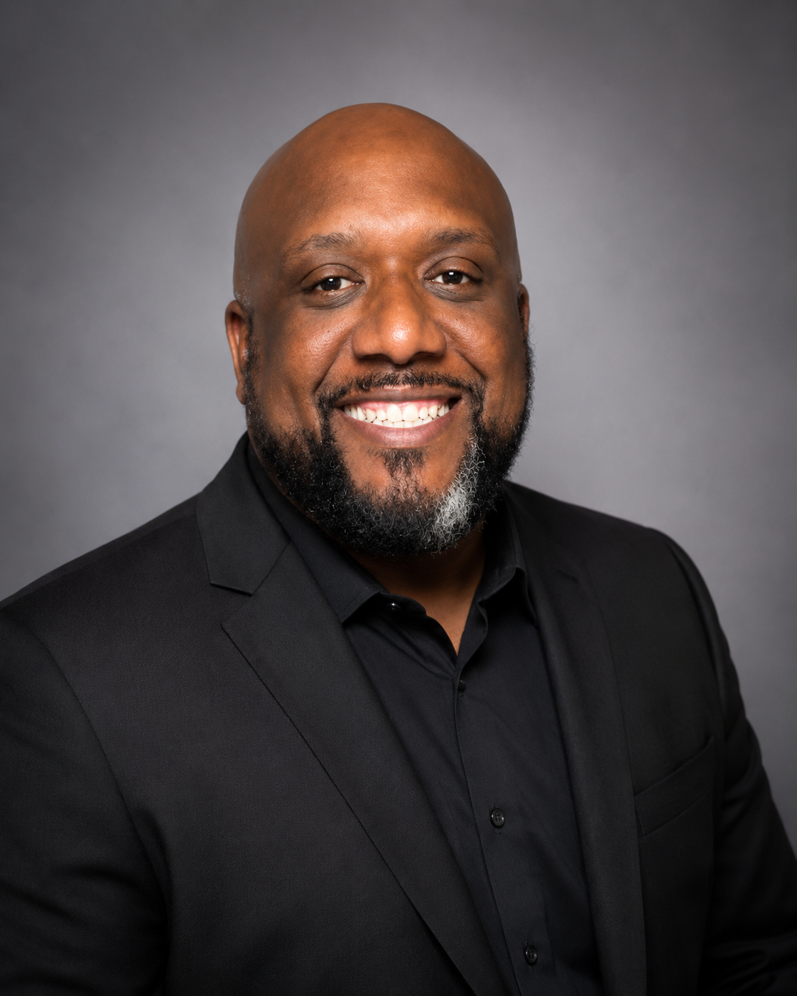 Portrait of a smiling Black man with a beard and shaved head, dressed in a black suit and shirt, against a gray background.
