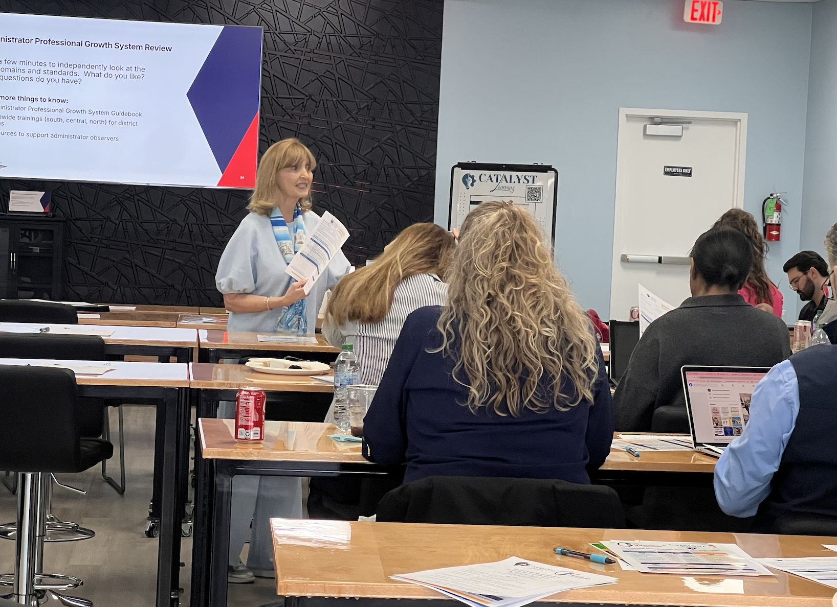 A woman standing and speaking in a conference room with a projected presentation titled 'Administrator Professional Growth System Review.' Several seated attendees are listening, some taking notes or using laptops. The room has a modern design with black patterned walls, and tables with papers, drinks, and laptops are visible. An exit door and fire extinguisher are in the background.