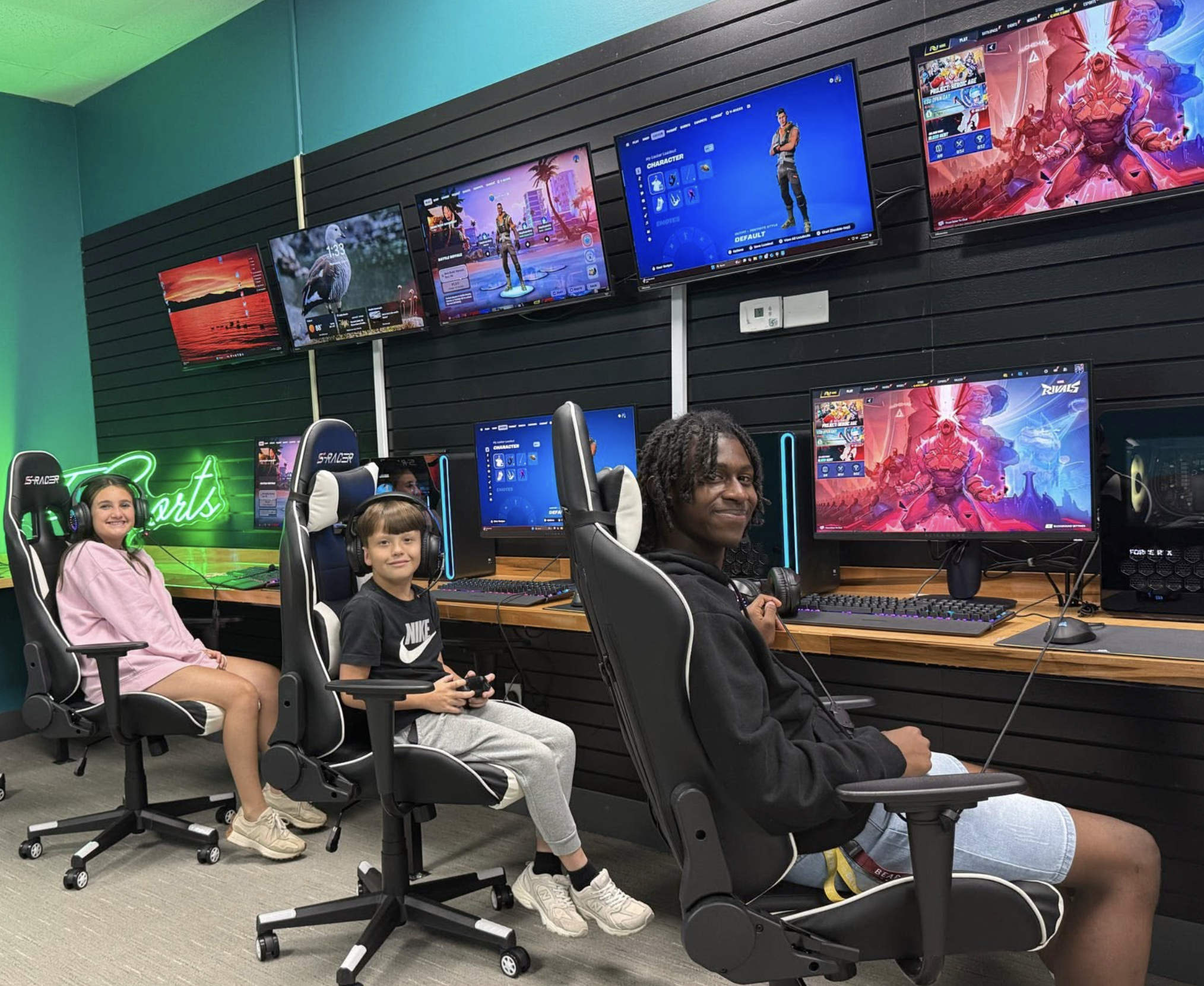 Three children and an adult in a gaming room with multiple monitors displaying video games. The children are seated at gaming stations, wearing headsets. The room has black slat wall paneling, and the monitors show colorful game scenes. There is a neon green sign reading 'Rivals' on the wall.
