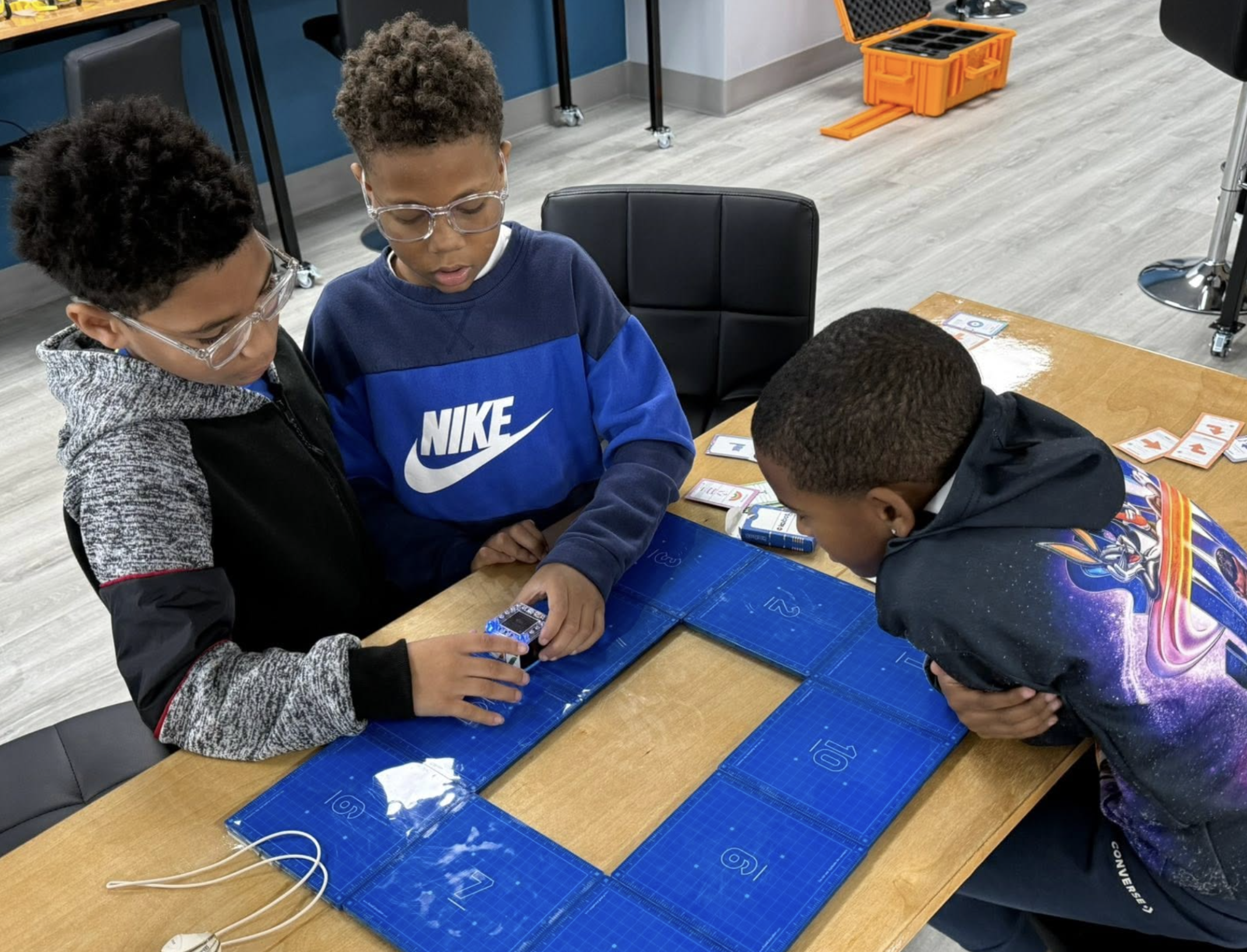 Four children gathered around a table, playing a card game with name tags and a blue gaming mat, in a classroom or game room setting.