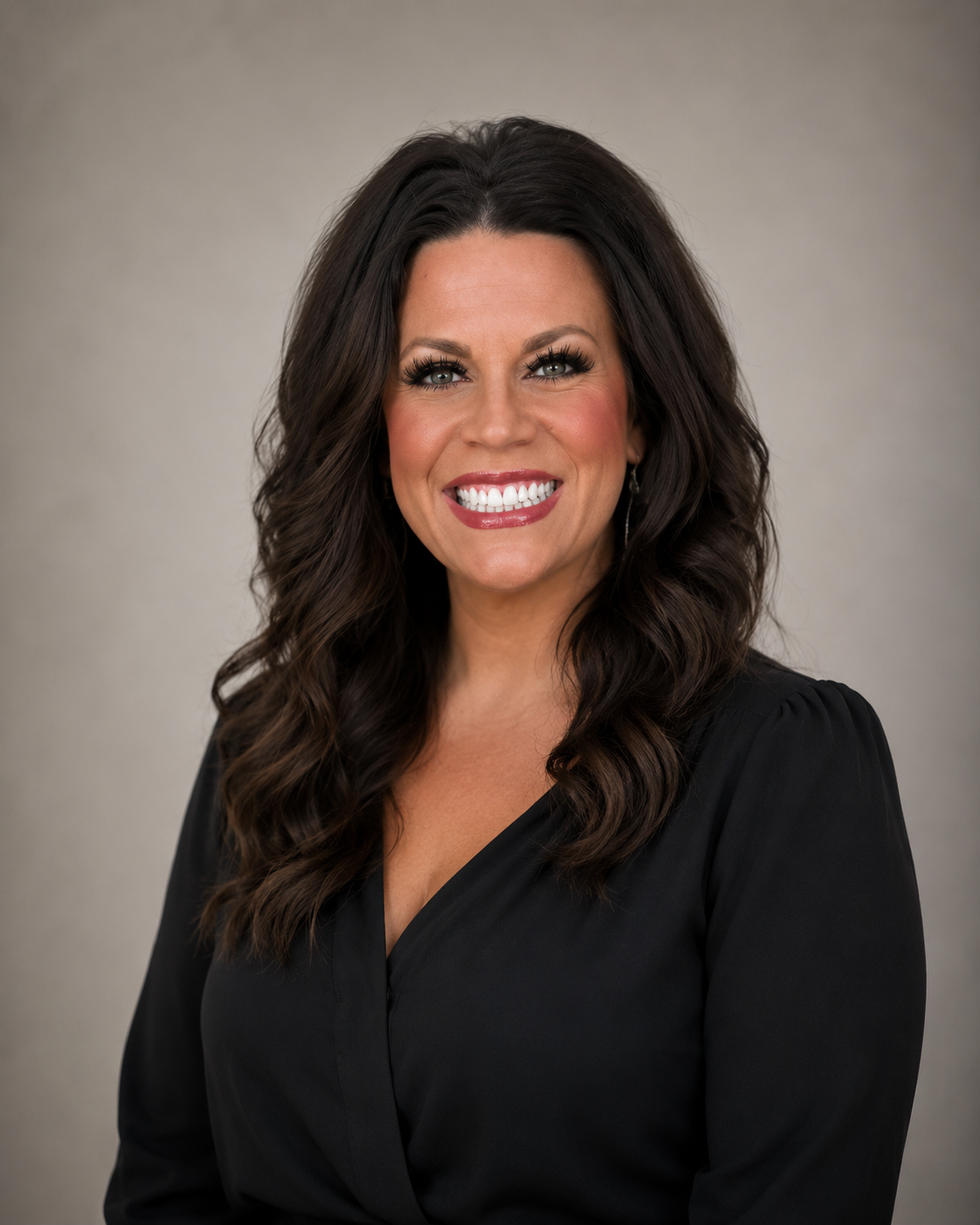 A woman with long, wavy dark hair and bright makeup, smiling and wearing a black top, posed against a neutral background.
