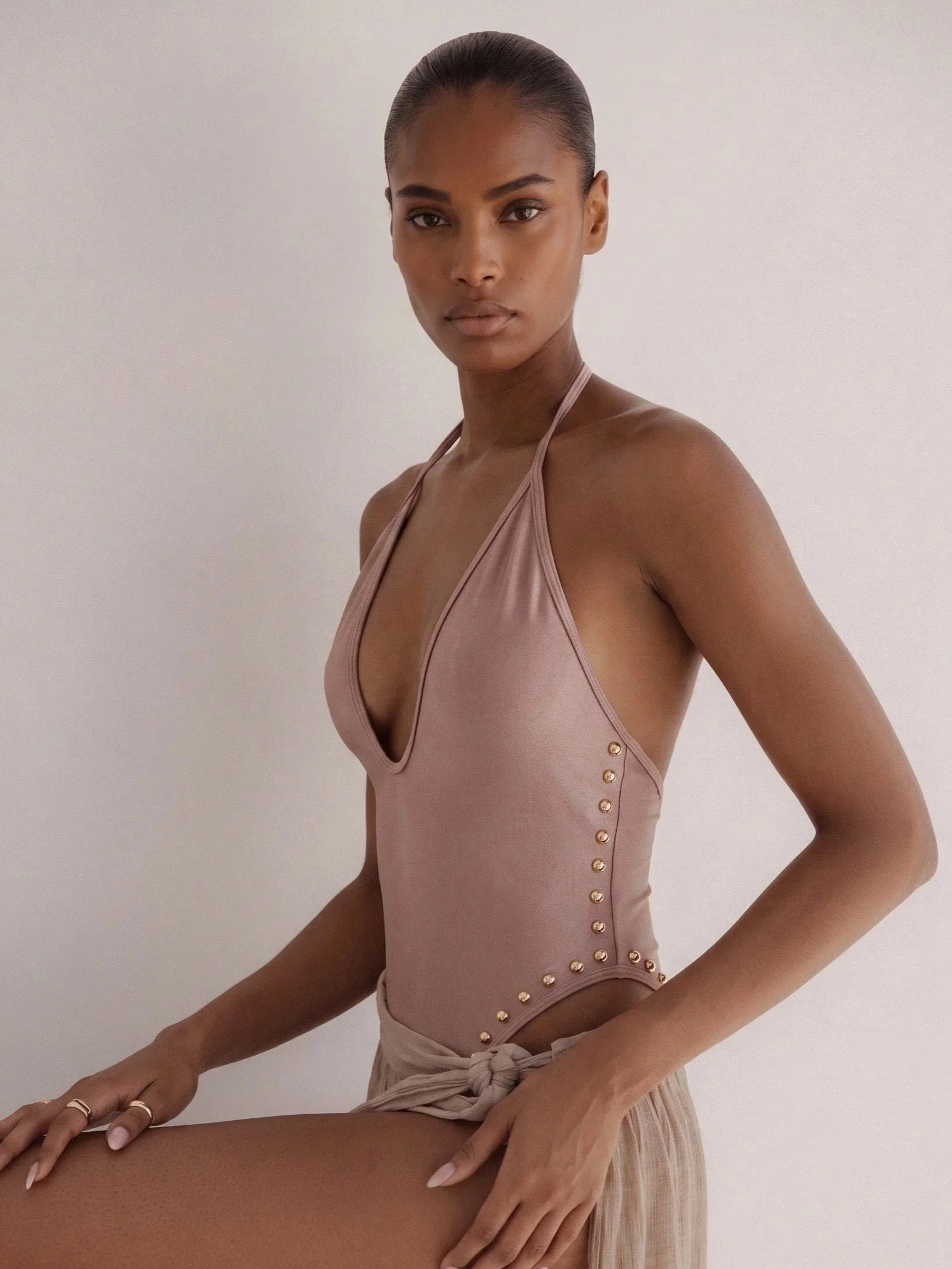 A woman with smooth dark skin and slicked-back hair posing against a plain white wall, wearing a pink halter top with gold button details along the side, and a light-colored skirt tied at the waist.