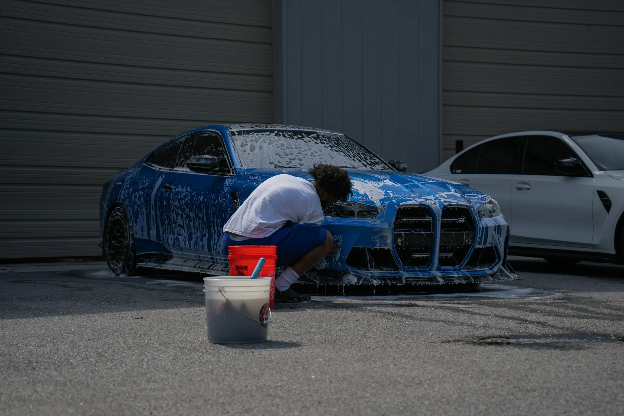 A person washing a blue sports car with soap and water outside. There are two buckets of cleaning supplies in the foreground, and a white car parked nearby. The background features a metal building with horizontal siding.