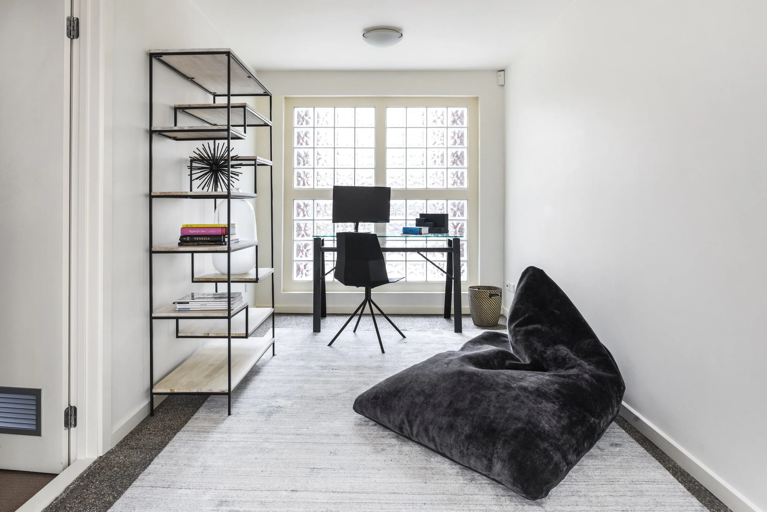A small home office with a black desk, black chair, glass top, and a computer monitor, set against a large window with frosted glass blocks, next to a black metal and wood shelving unit with decorative items and books, and a black plush armchair in the foreground.