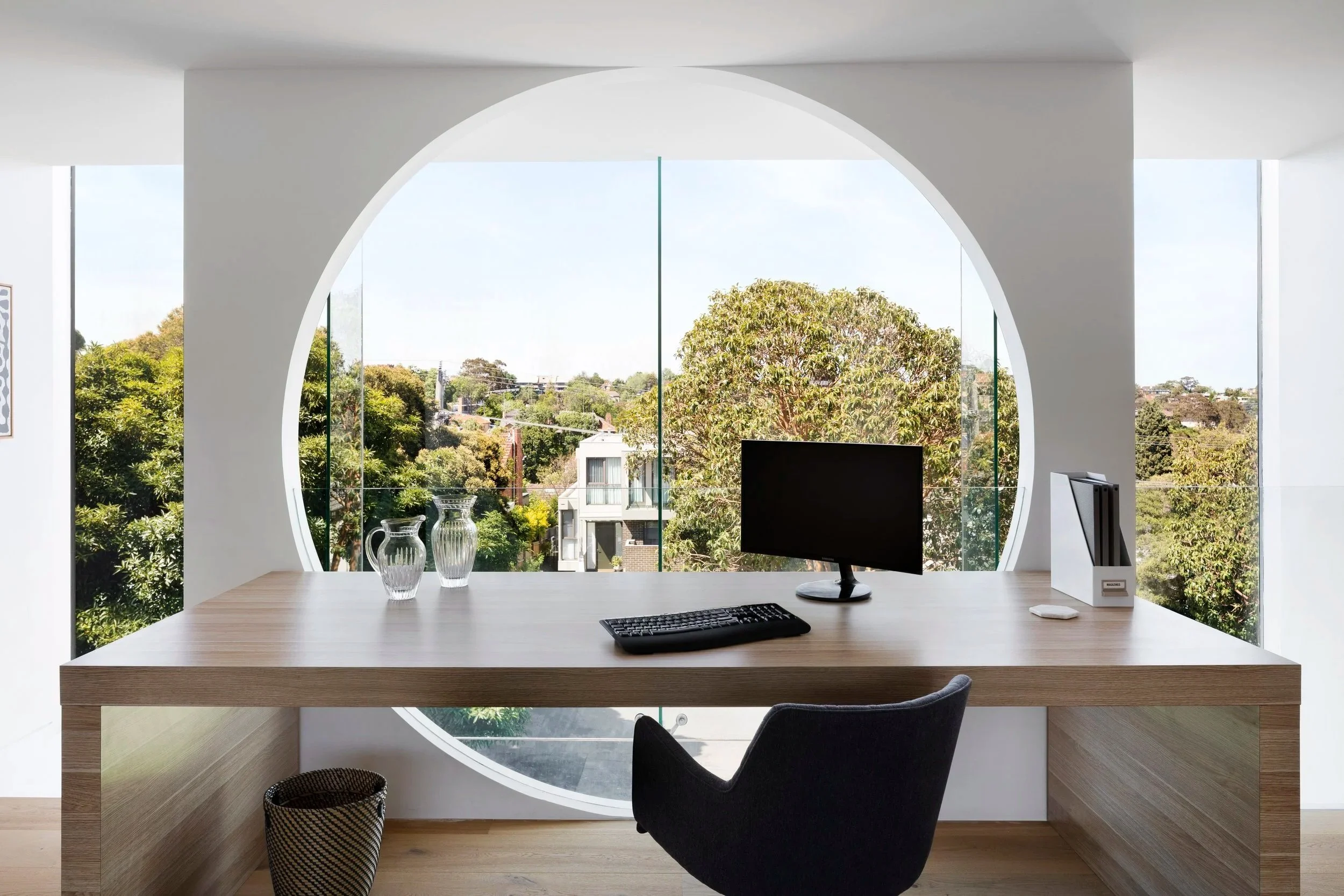 Modern minimalist office with a large circular window, a wooden desk, a computer monitor, a keyboard, a mouse, and a file organizer, overlooking a green outdoor view with trees and houses.