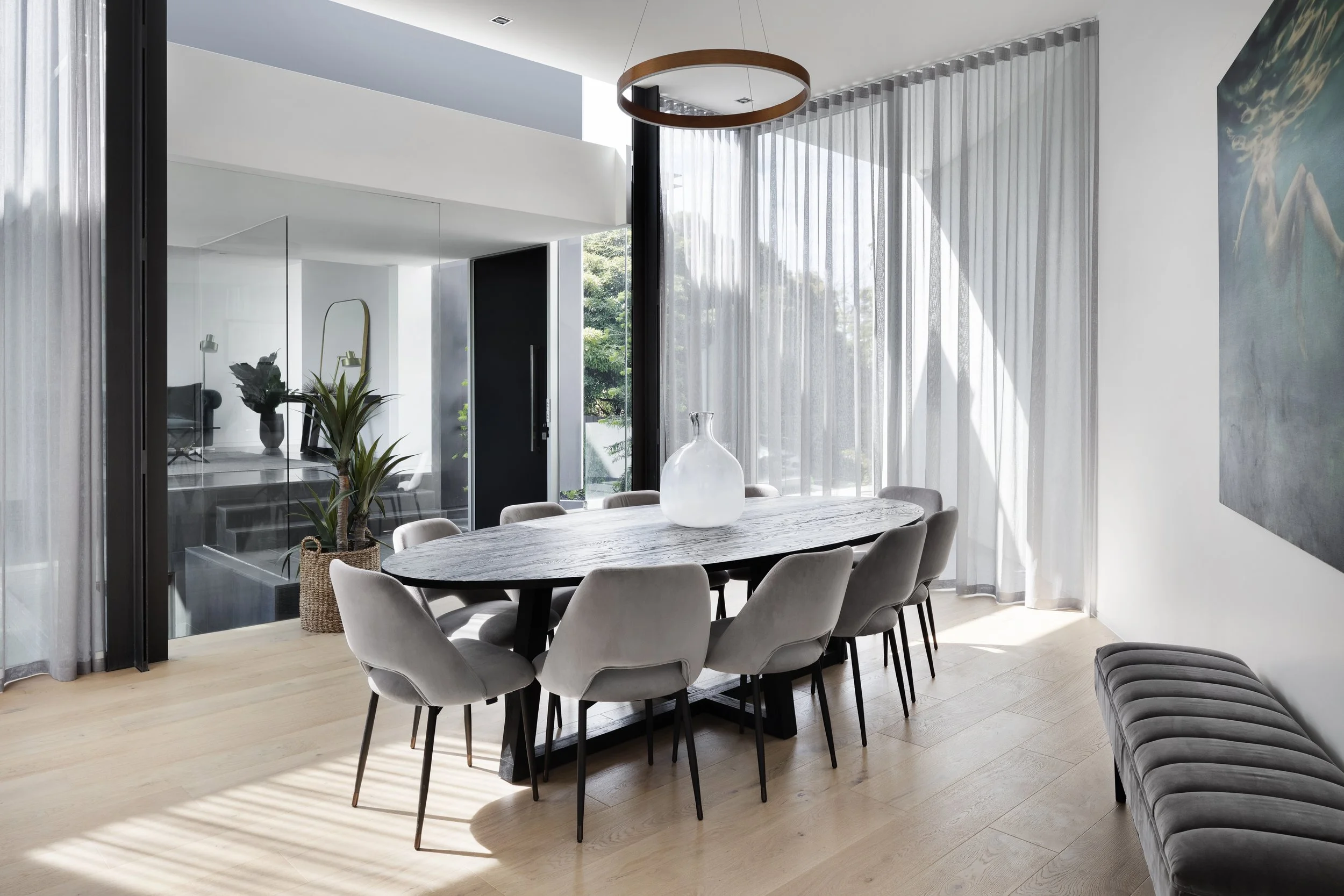 Modern dining room with a marble table, eleven beige upholstered chairs, a large glass vase, and a side view of a gray cushioned bench. There are white curtains, a dark door, and a large painting with blue and green tones. Sunlight creates striped shadows on the light wood flooring.
