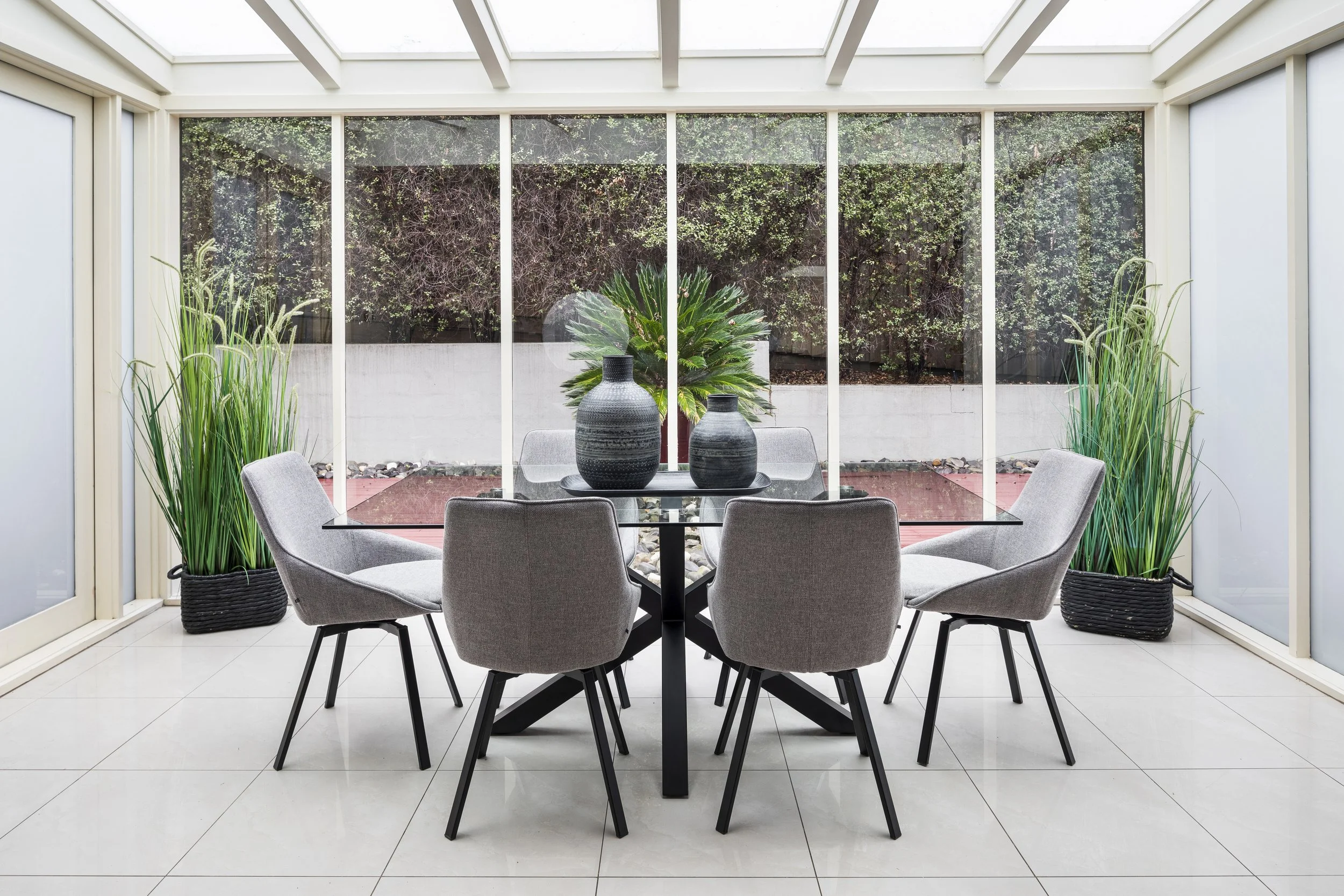 A bright dining room with a round glass table, six gray upholstered chairs, and two tall potted plants on either side. A large window with a view of a garden with trees and bushes.