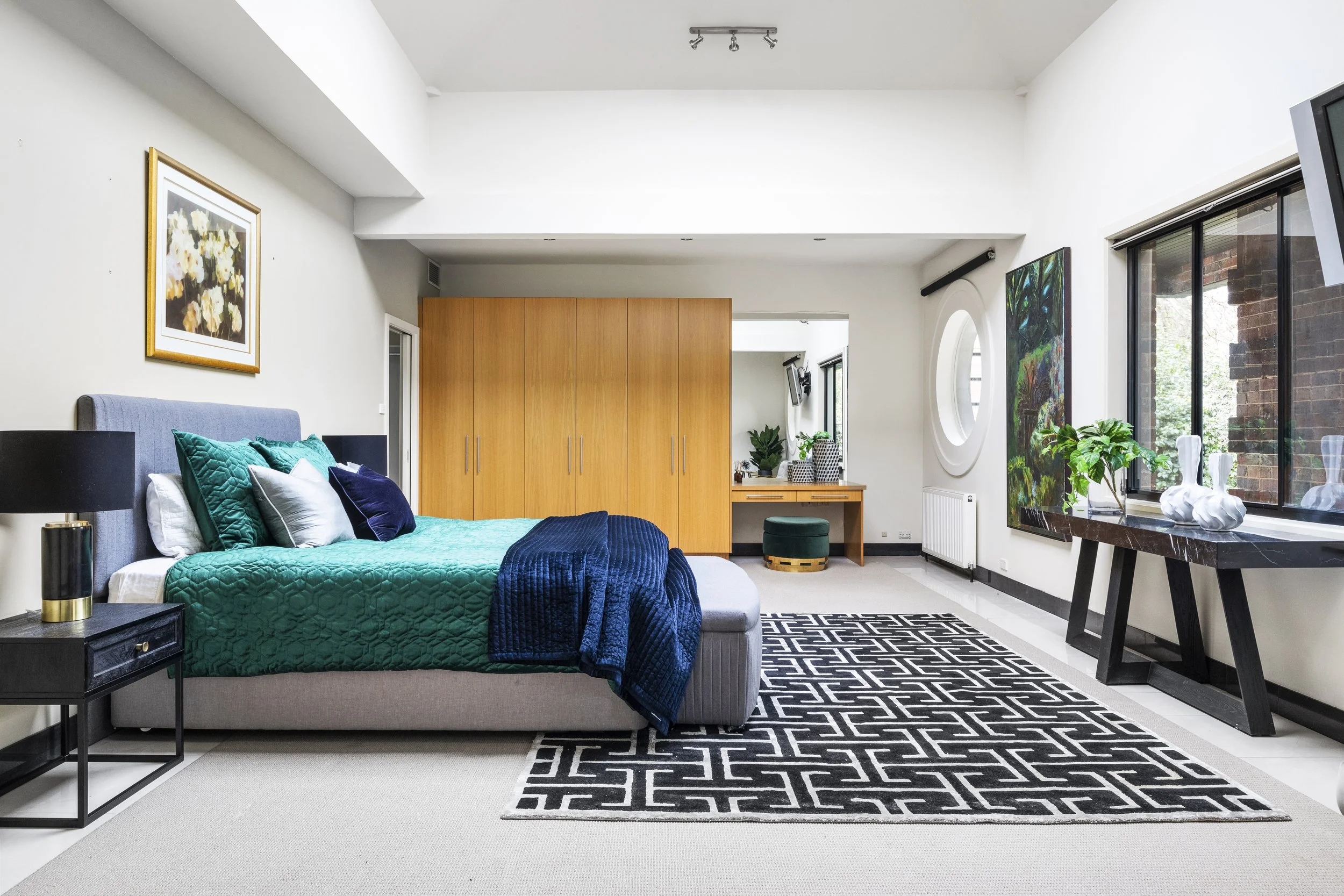 Modern bedroom featuring a gray bed with teal and white pillows and a dark blue blanket, black nightstand, large black and white geometric rug, white walls with framed floral artwork, wooden wardrobe, round window, colorful art piece, glass sofa view outside, and a black table with white sculptures.