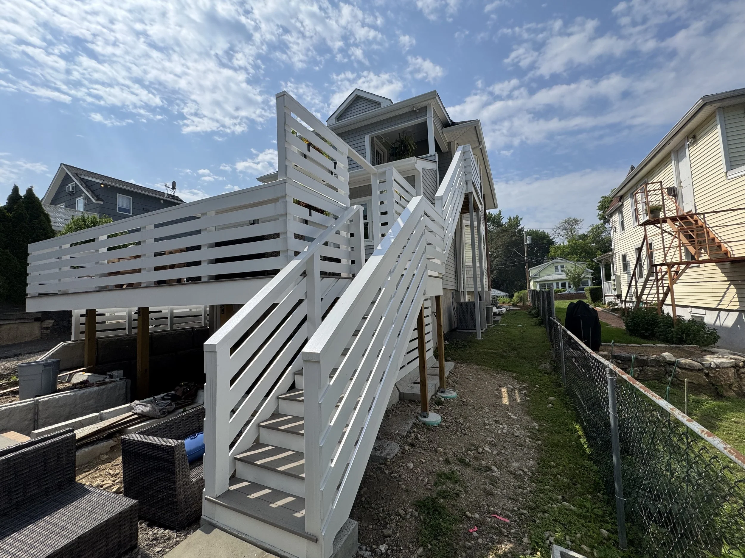 White exterior staircase leading to the second floor of a house under construction, with neighboring houses and a partly cloudy sky in the background.