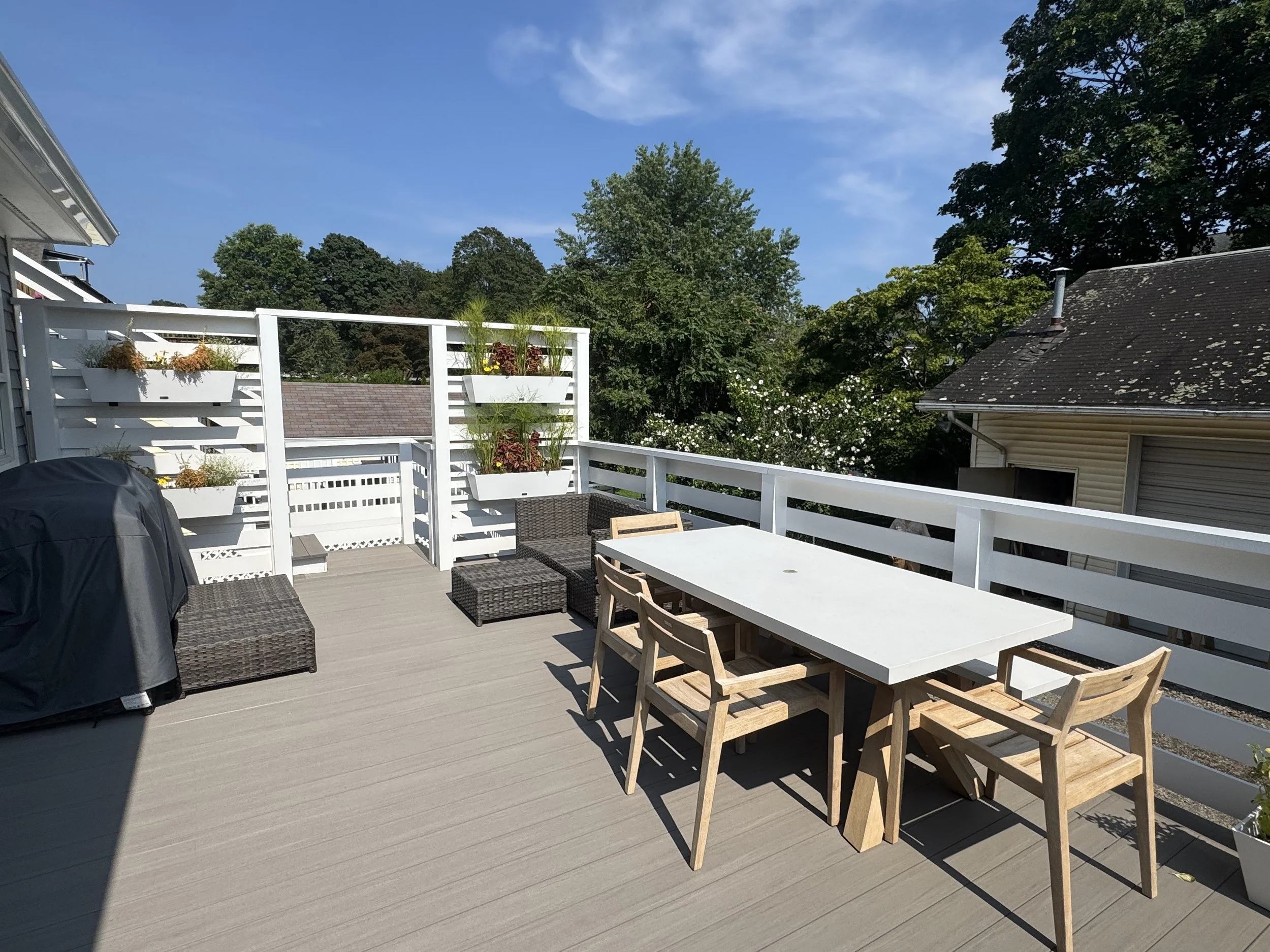 A spacious outdoor balcony with lounge chairs, a wooden dining table, and white planters with flowers, surrounded by trees and neighboring houses, under a clear blue sky.